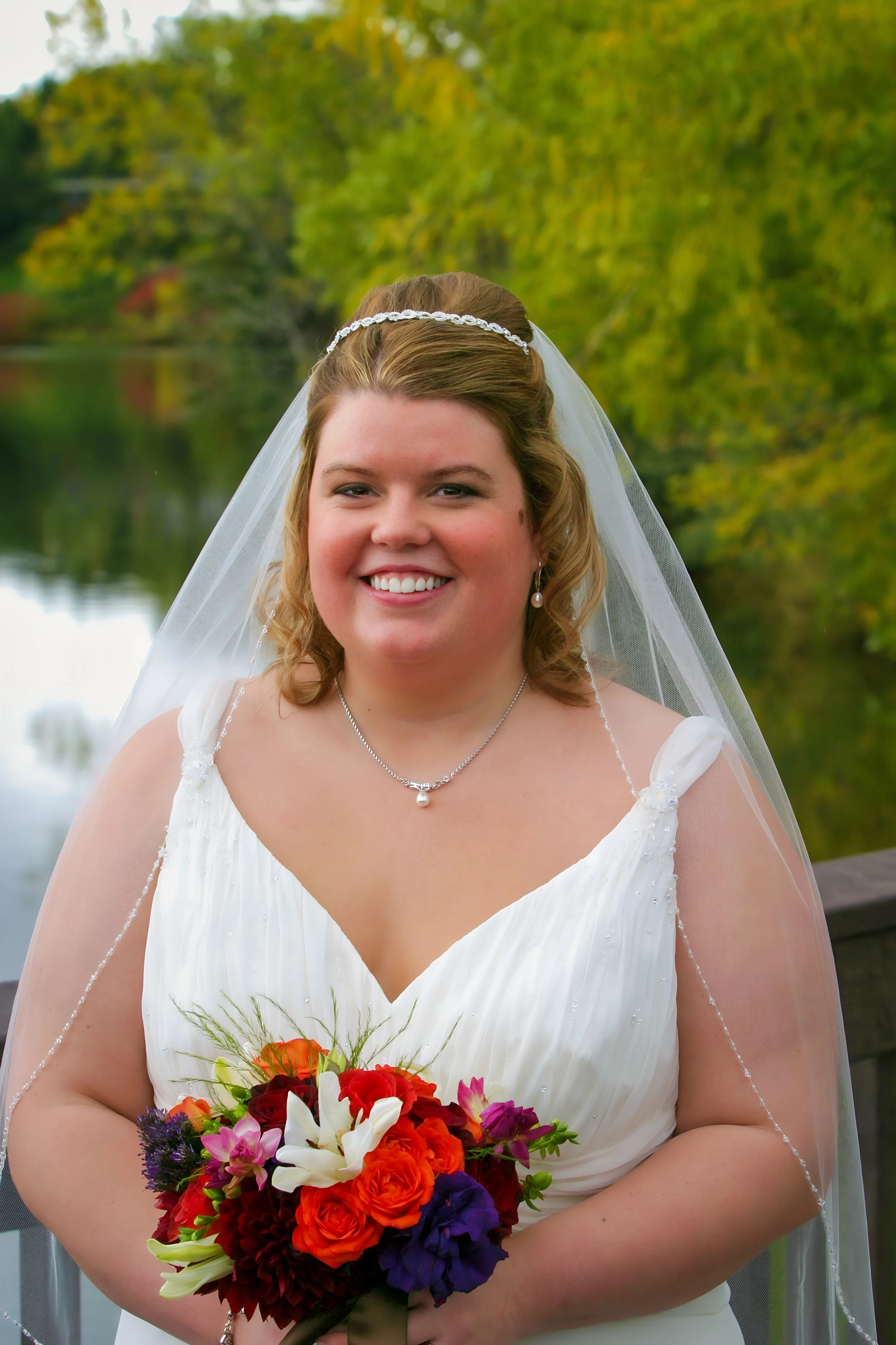 A smiling woman dressed as a bride, holding a colorful bouquet of flowers, standing outdoors near a body of water with green trees in the background.