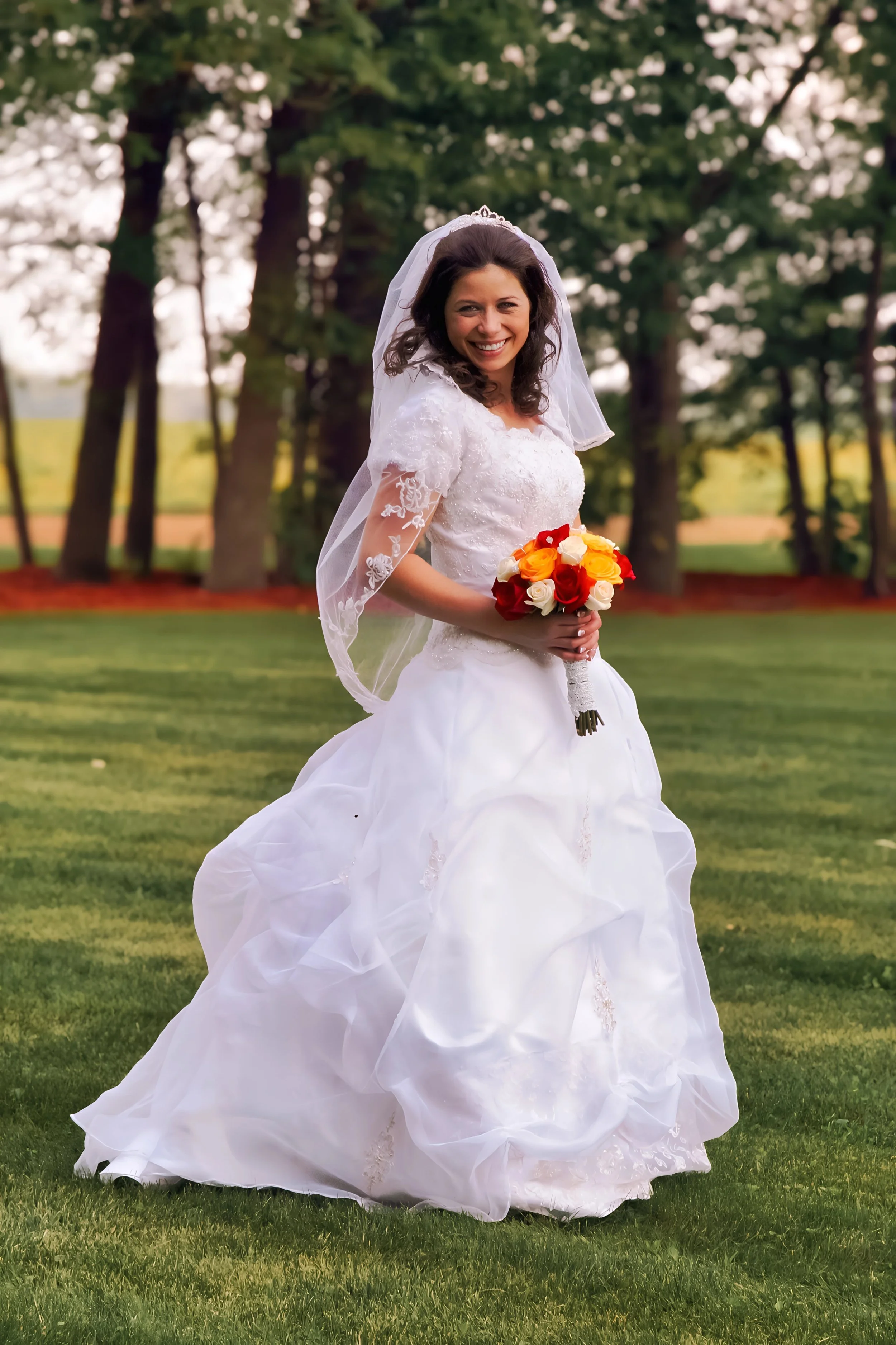 A bride in a white wedding dress holding a colorful bouquet of red, orange, and white roses, smiling outdoors on a grassy area with trees in the background.