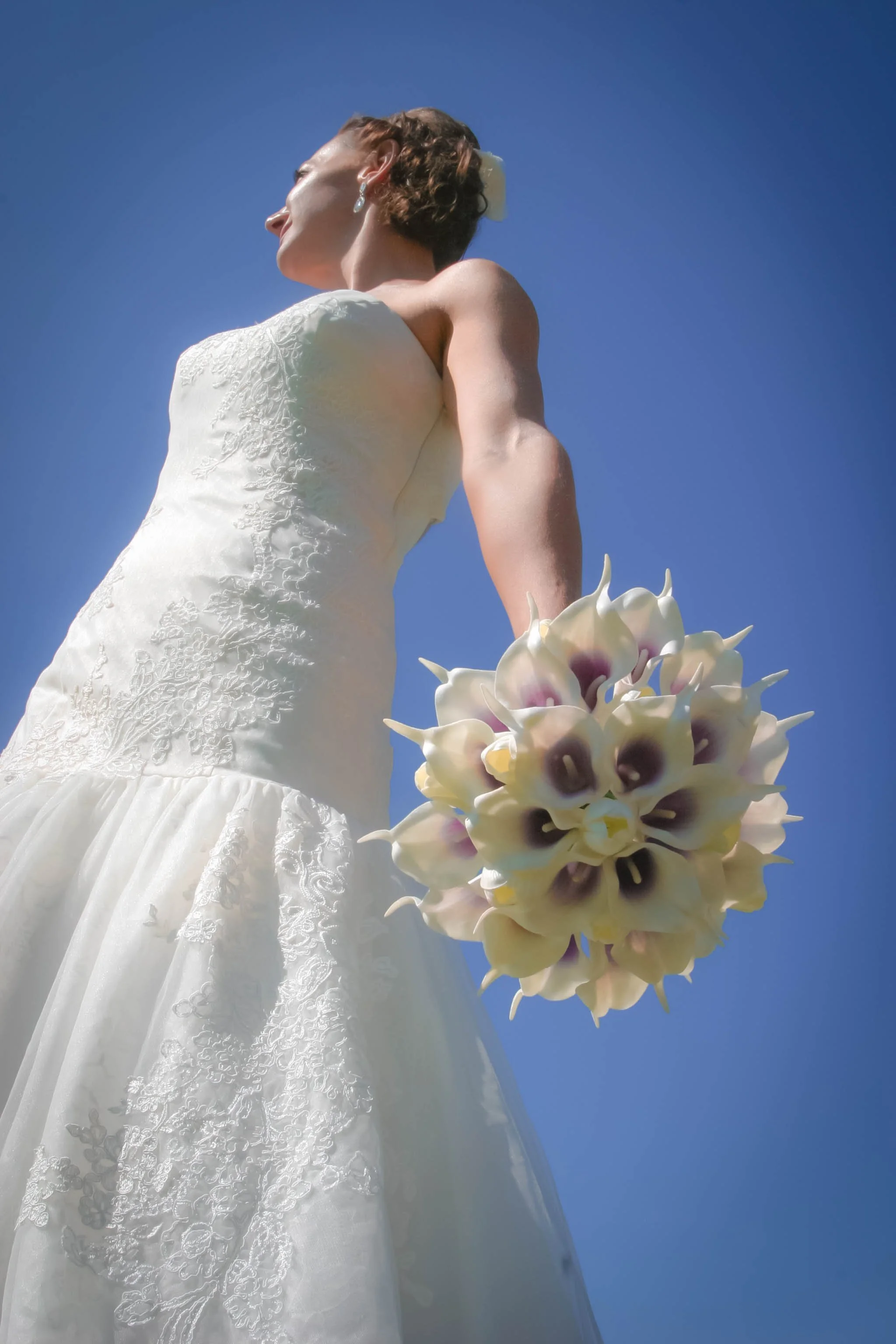 A woman in a white wedding dress holding a bouquet of white and purple flowers, standing outdoors against a clear blue sky.
