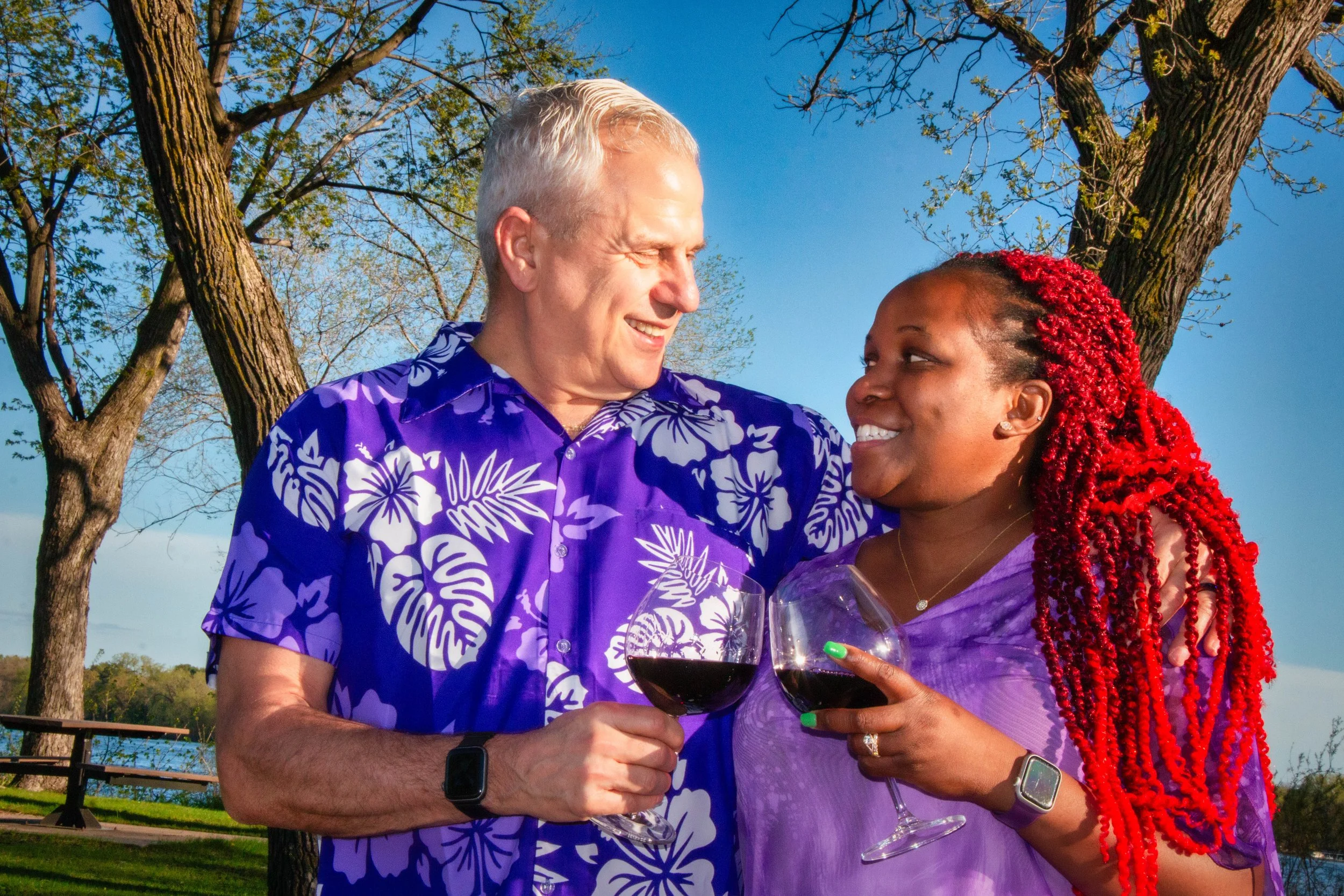 A smiling elderly man and woman with red dreadlocks clink glasses of red wine outdoors on a sunny day, with trees and blue sky in the background.