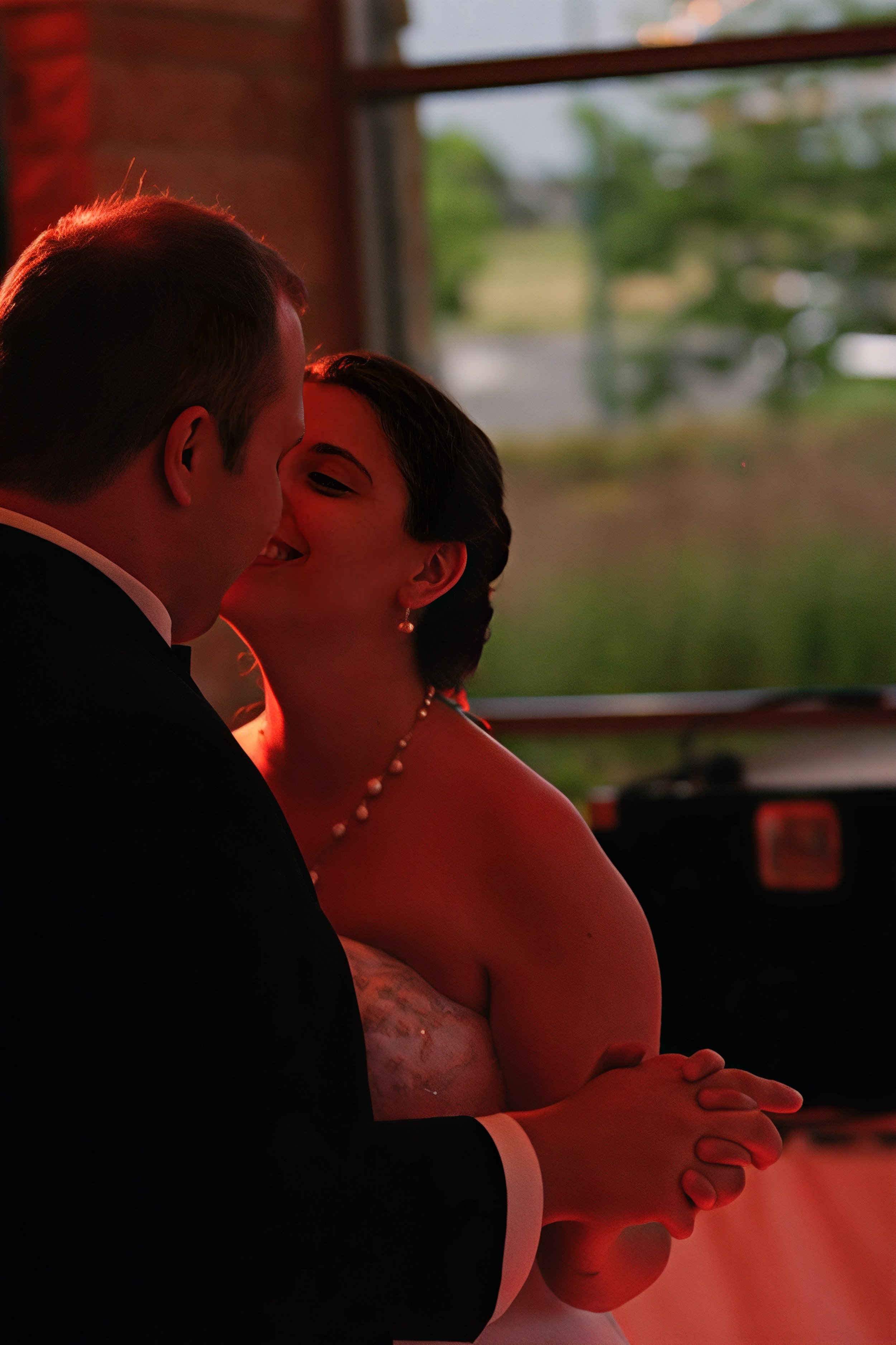 A bride and groom share a kiss during a wedding reception, with the bride smiling and wearing a pearl necklace, near a large window showing a blurred outdoor landscape.
