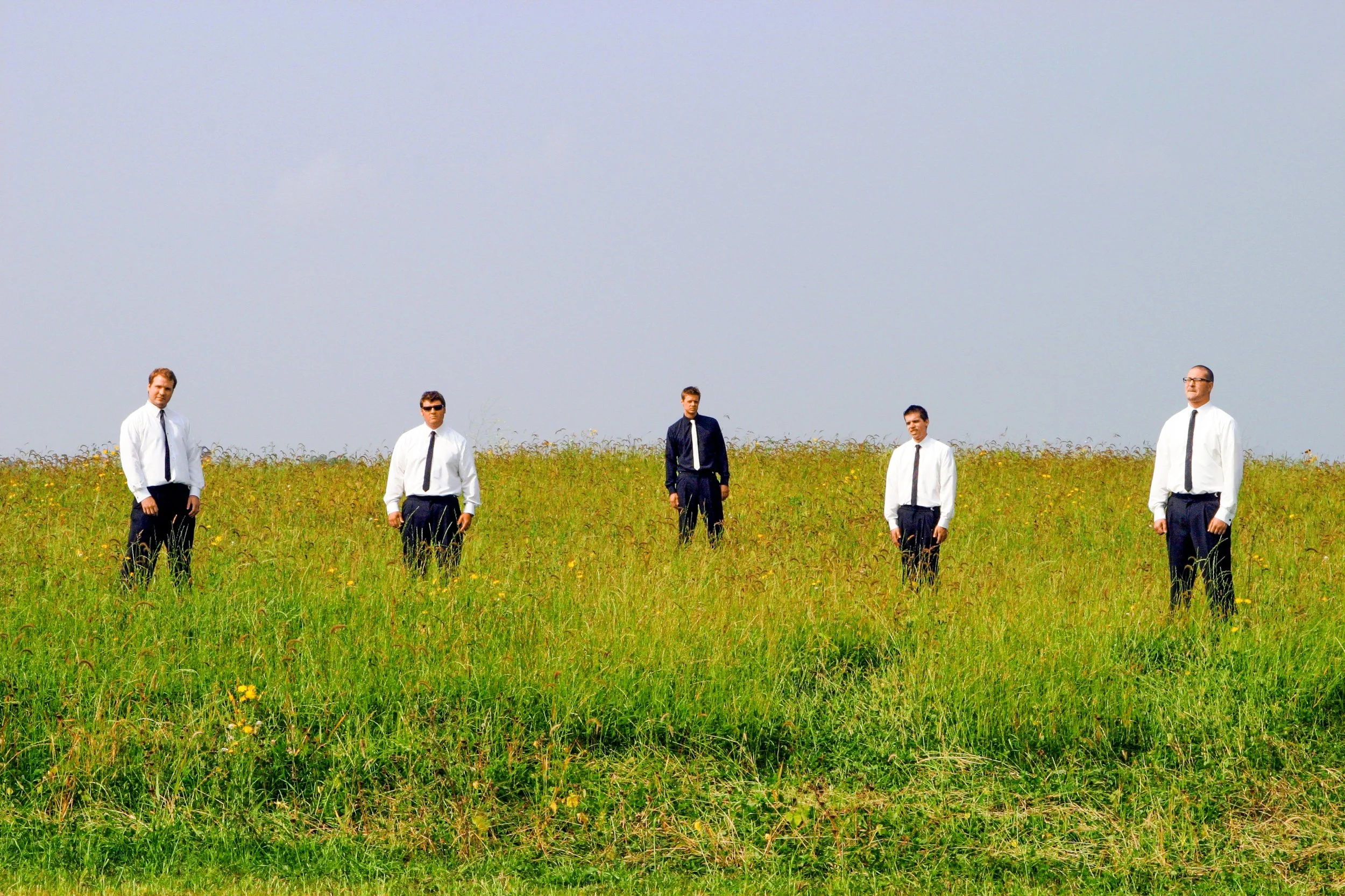 Five men in formal business attire standing in a grassy field with a cloudy sky background.