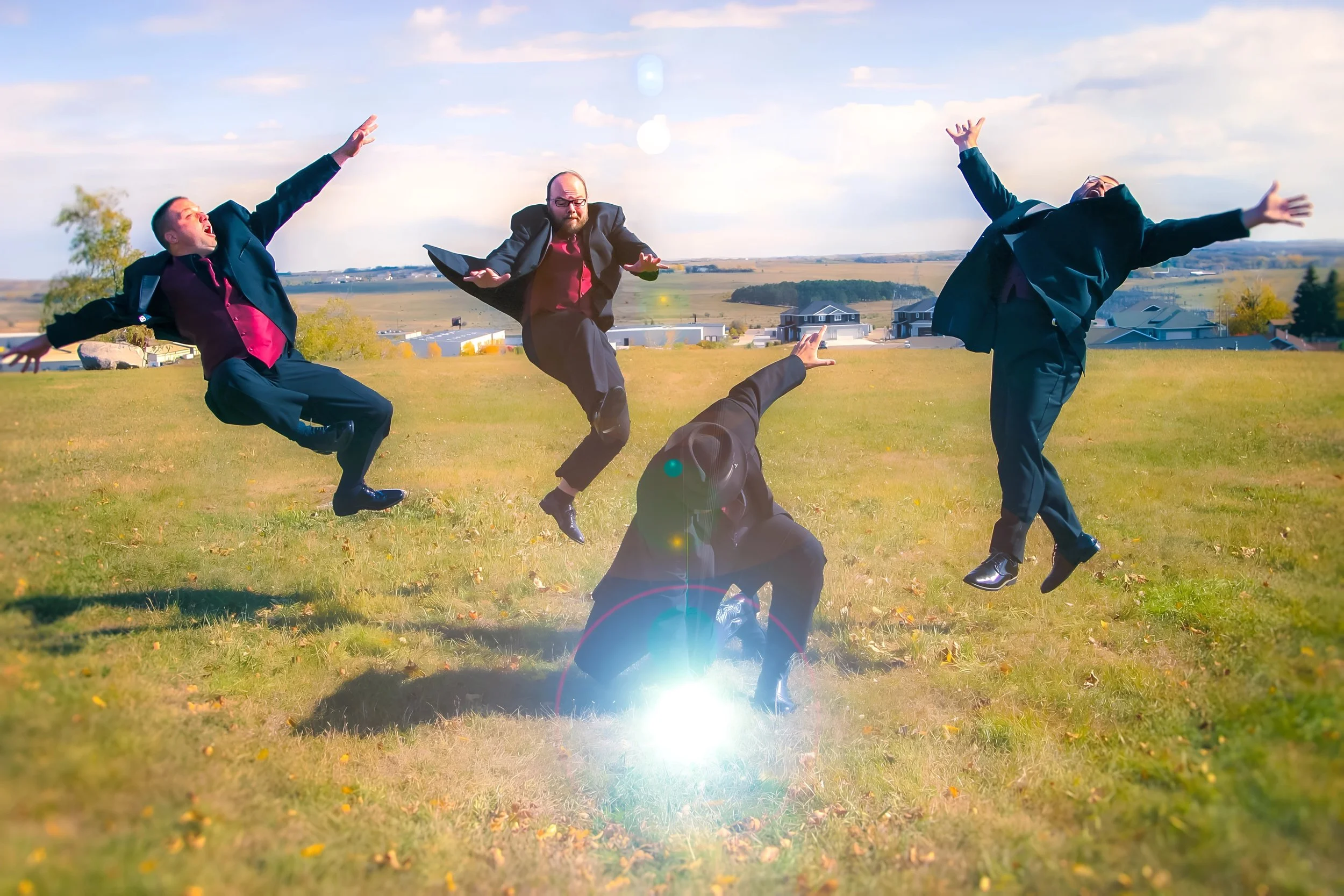Four men in suits jumping in the air on a grassy field with a clear sky and houses in the background.