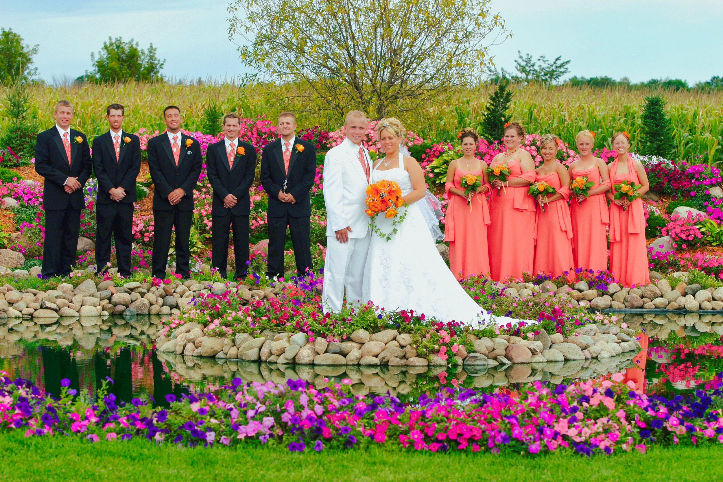 Wedding party outdoors with bride and groom in the center, six groomsmen in dark suits to the left, and seven bridesmaids in coral dresses to the right, all holding bouquets, surrounded by colorful flowers and a pond.