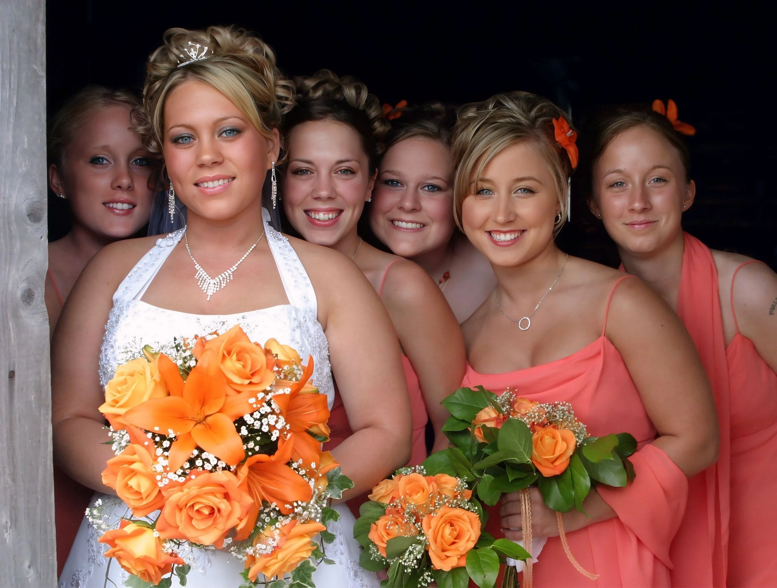A group of six women dressed in peach-colored dresses, holding orange bouquets of roses and lilies, smiling for a photo.
