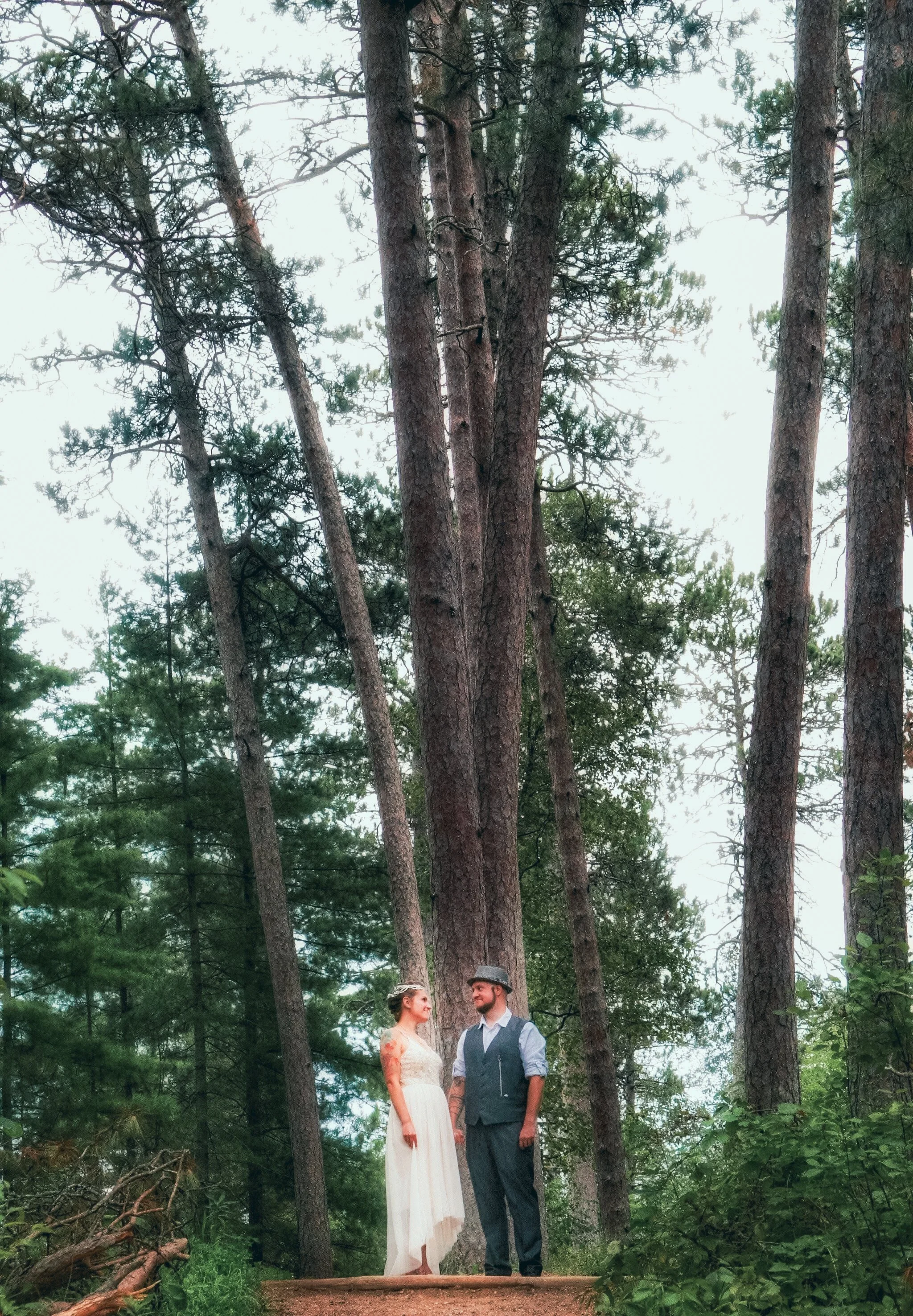 A man and woman dressed in vintage wedding attire standing on a dirt path in a forest with tall pine trees and green foliage.