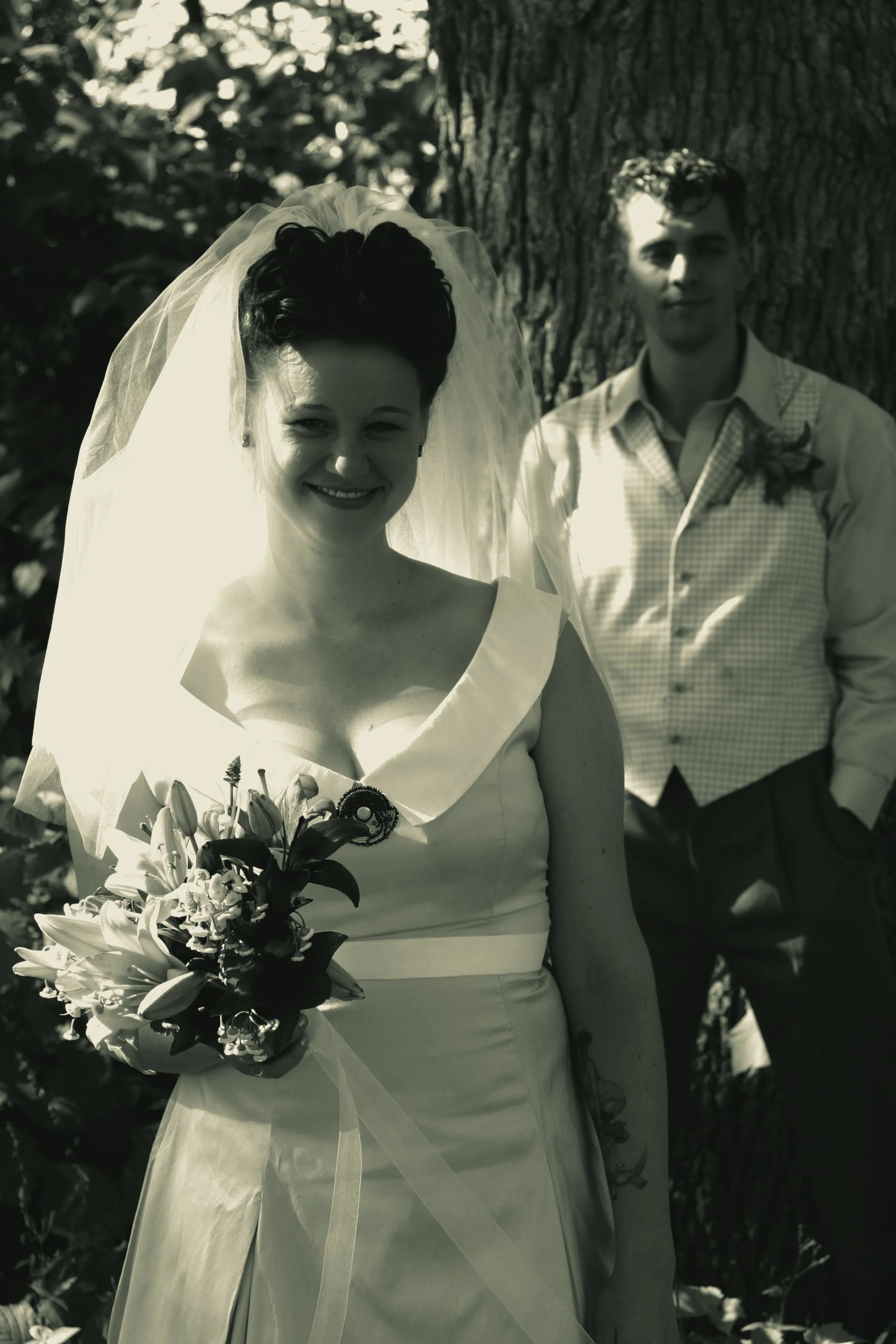 Black and white photo of a bride smiling, holding a bouquet of flowers, standing outdoors near a tree, with a groom in the background leaning against the tree.