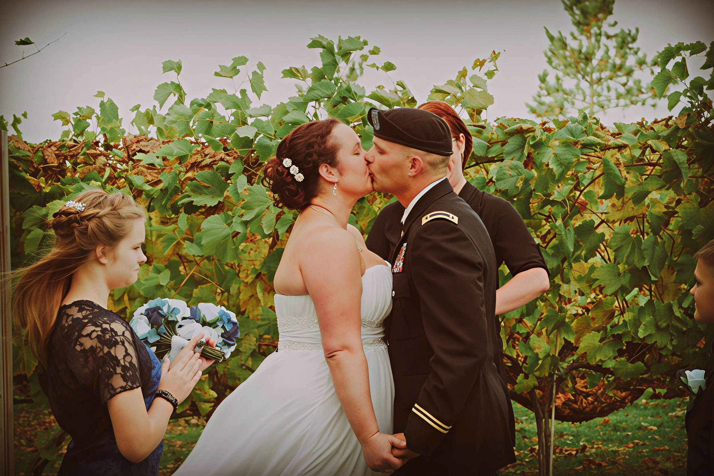 A bride and a groom in uniform share a kiss during their wedding ceremony outdoors, surrounded by green foliage. Two young girls are present, one holding a ring pillow and the other watching the couple.