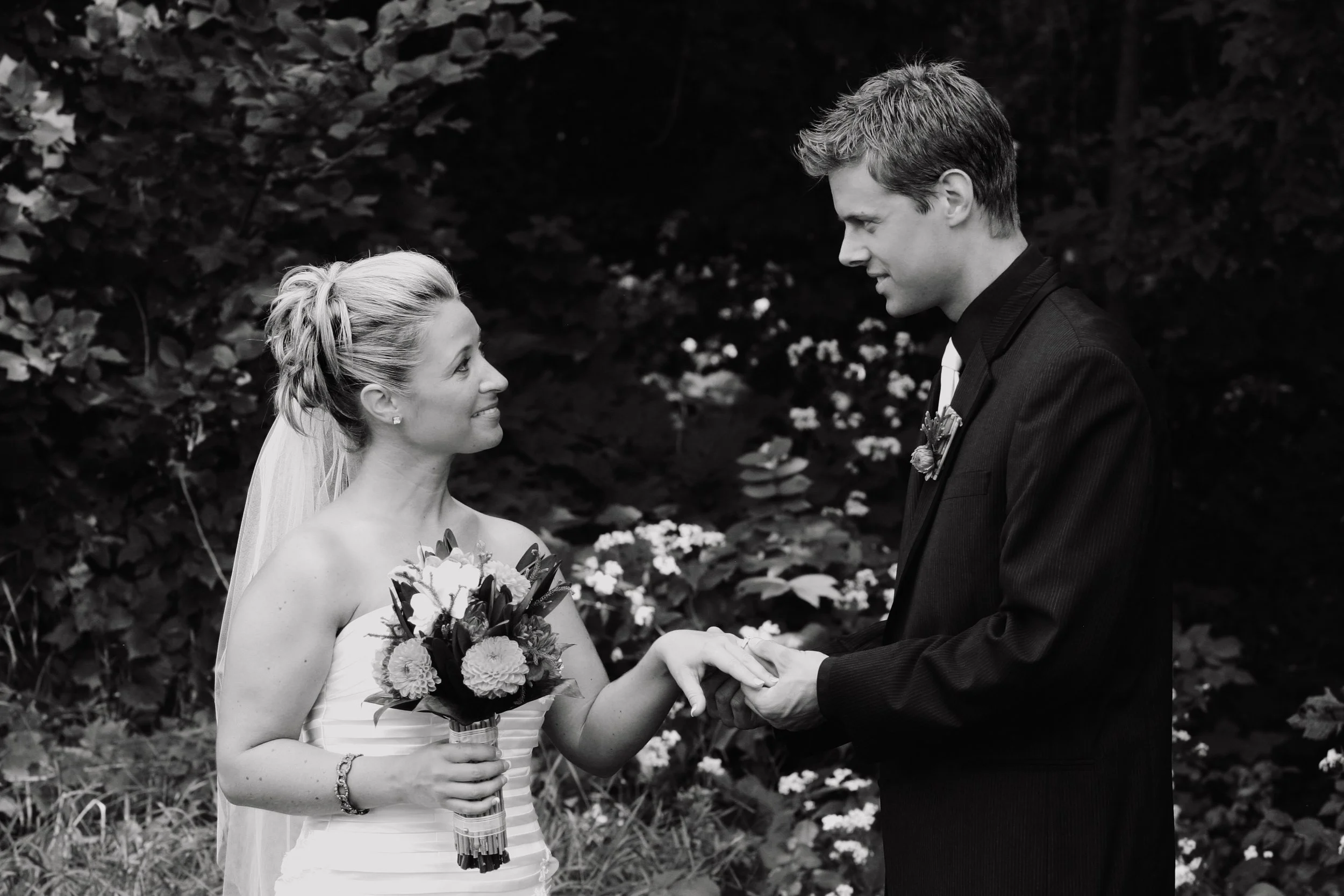 A black and white photo of a bride and groom exchanging rings outdoors, with the bride holding a bouquet and smiling at the groom.