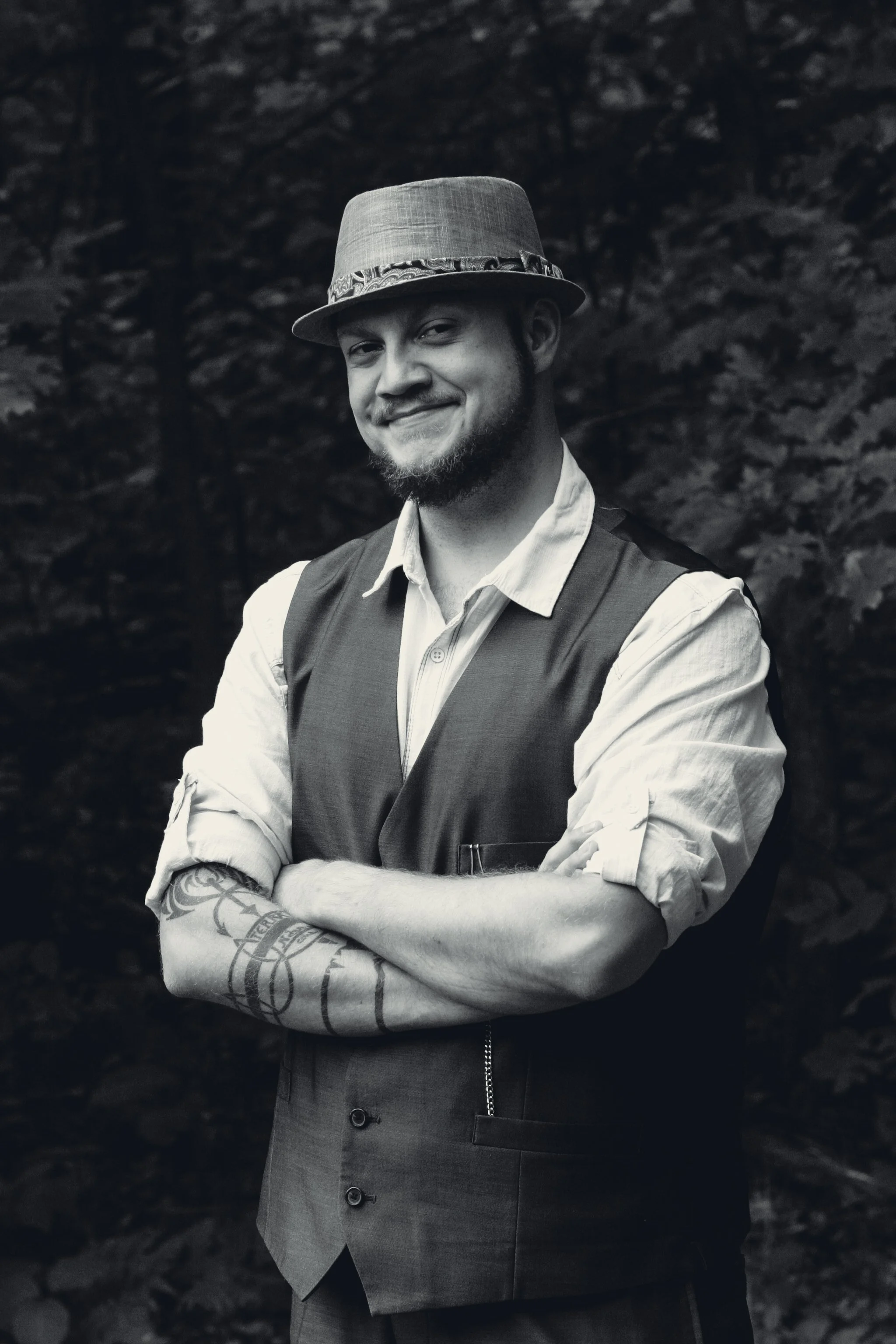 A man with a beard and mustache wearing a fedora hat, a vest over a button-up shirt with rolled-up sleeves, standing outdoors with arms crossed and smiling in black and white.