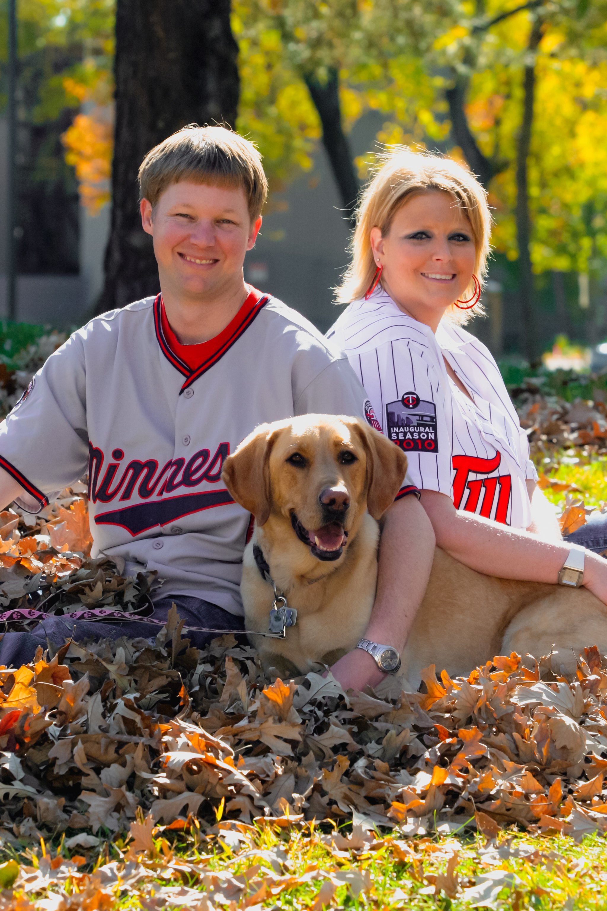 A smiling couple and their dog sitting on autumn leaves in a park.