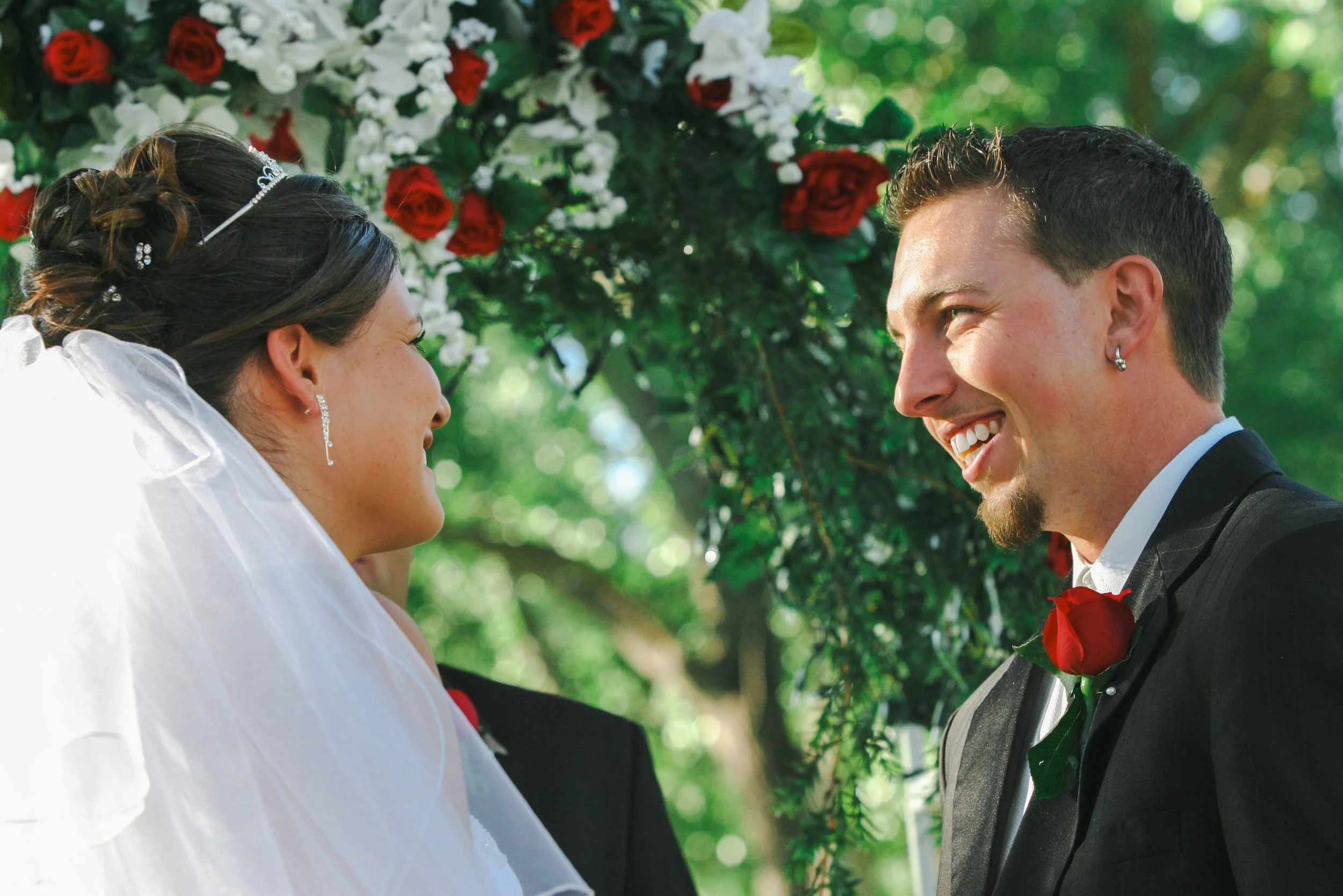 A bride and groom face each other smiling during a wedding ceremony outdoors under a floral arch with red and white flowers and green foliage.