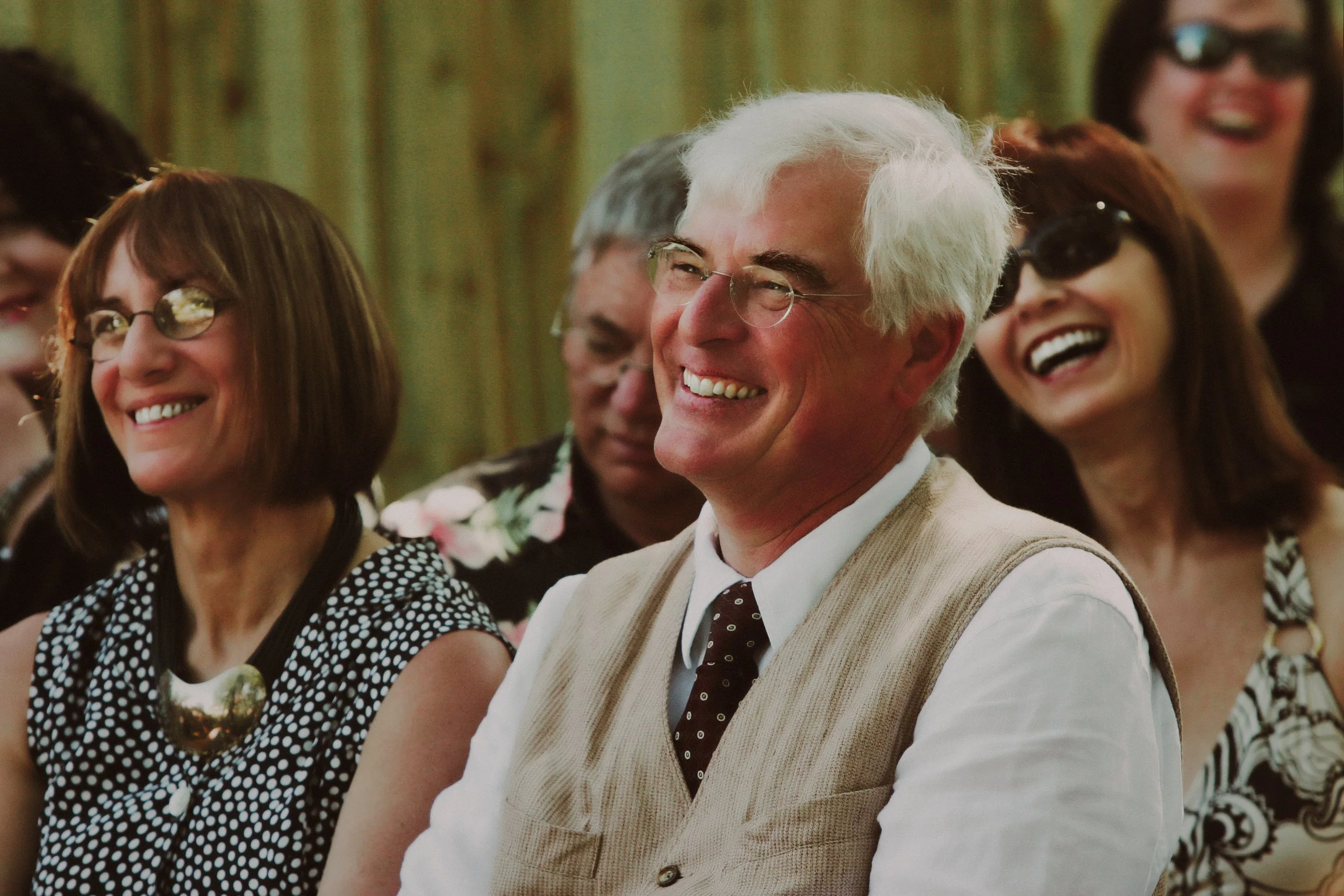 A group of people smiling and laughing at an outdoor event, with a man in the center wearing glasses, a white shirt, a beige vest, and a dark tie, surrounded by women wearing glasses and patterned clothing.
