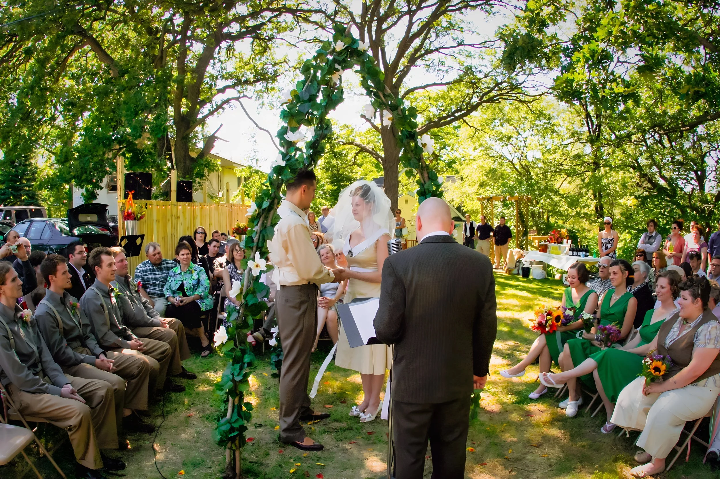 An outdoor wedding ceremony with a couple holding hands under a decorative arch surrounded by guests seated on either side, with lush trees and sunlight in the background.