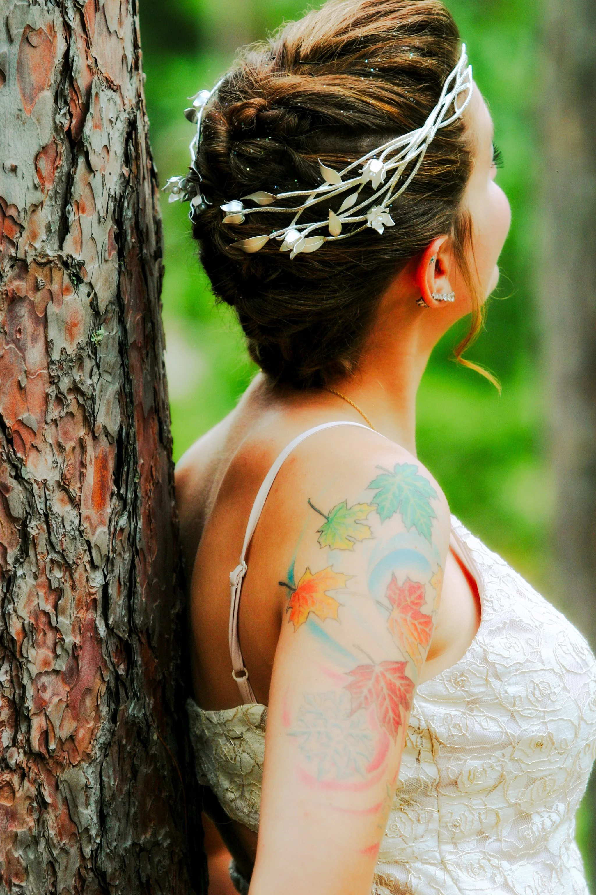 A woman with a updo hairstyle wearing a white dress, silver jewelry, and a white floral hair accessory, standing outdoors beside a tree, with colorful leaf tattoos on her shoulder and arm, against a blurred green background.