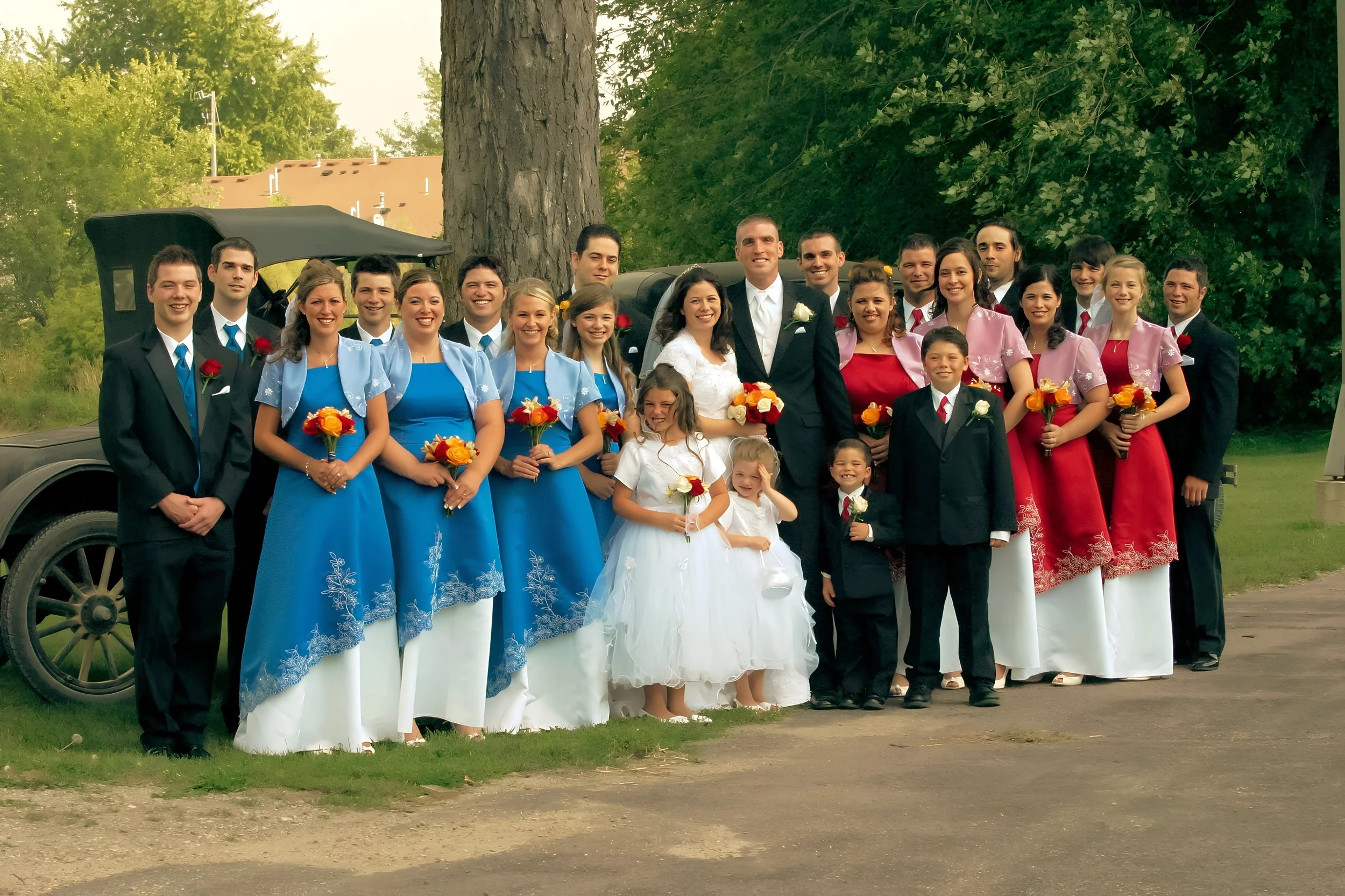 A wedding party outdoors with the bride and groom in the center, surrounded by bridesmaids in blue and pink dresses, and children in white and black attire, all posing for a group photo near a large tree and vintage car.
