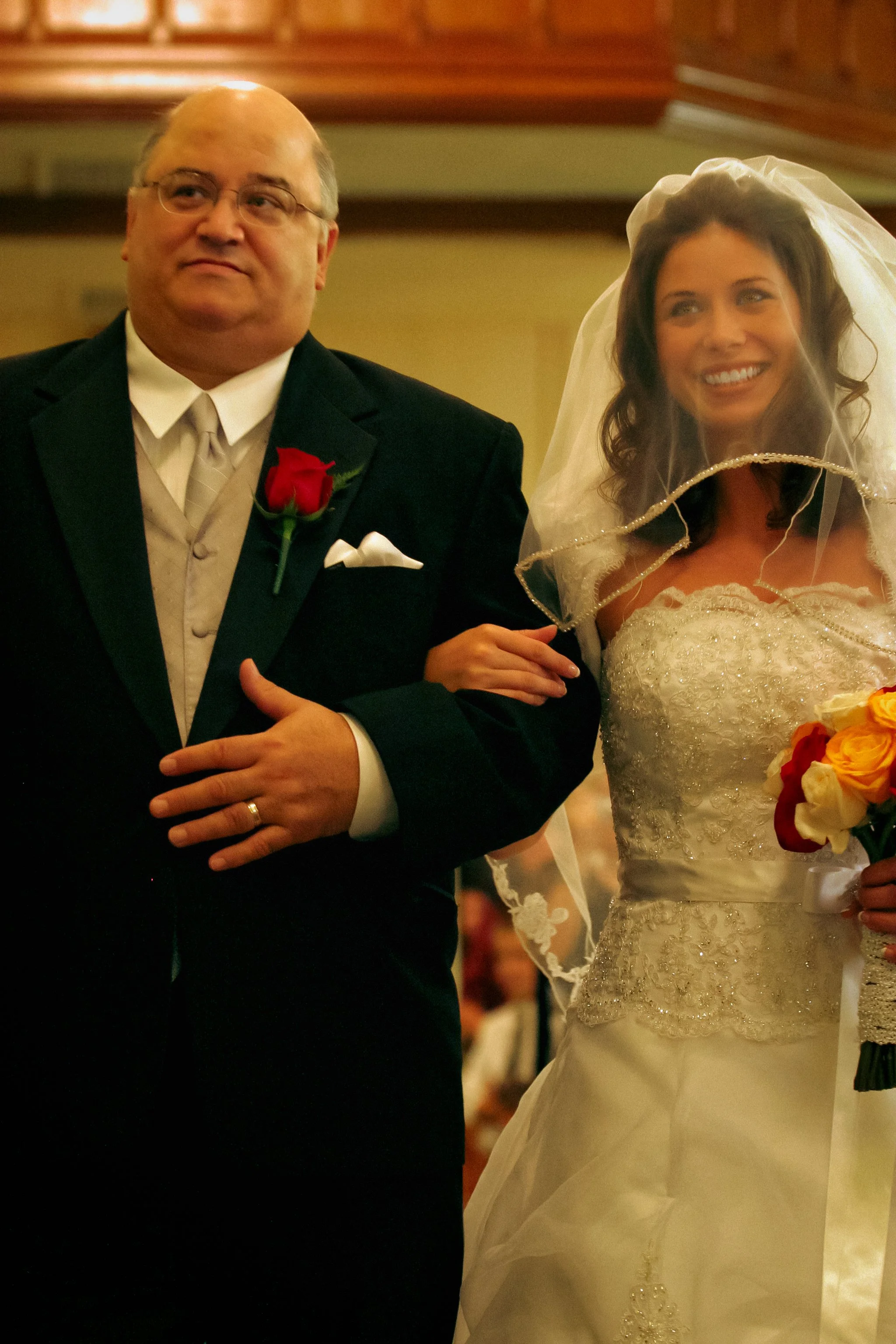 A bride in a white wedding dress and veil walking down the aisle with a man in a black suit, in a church setting.