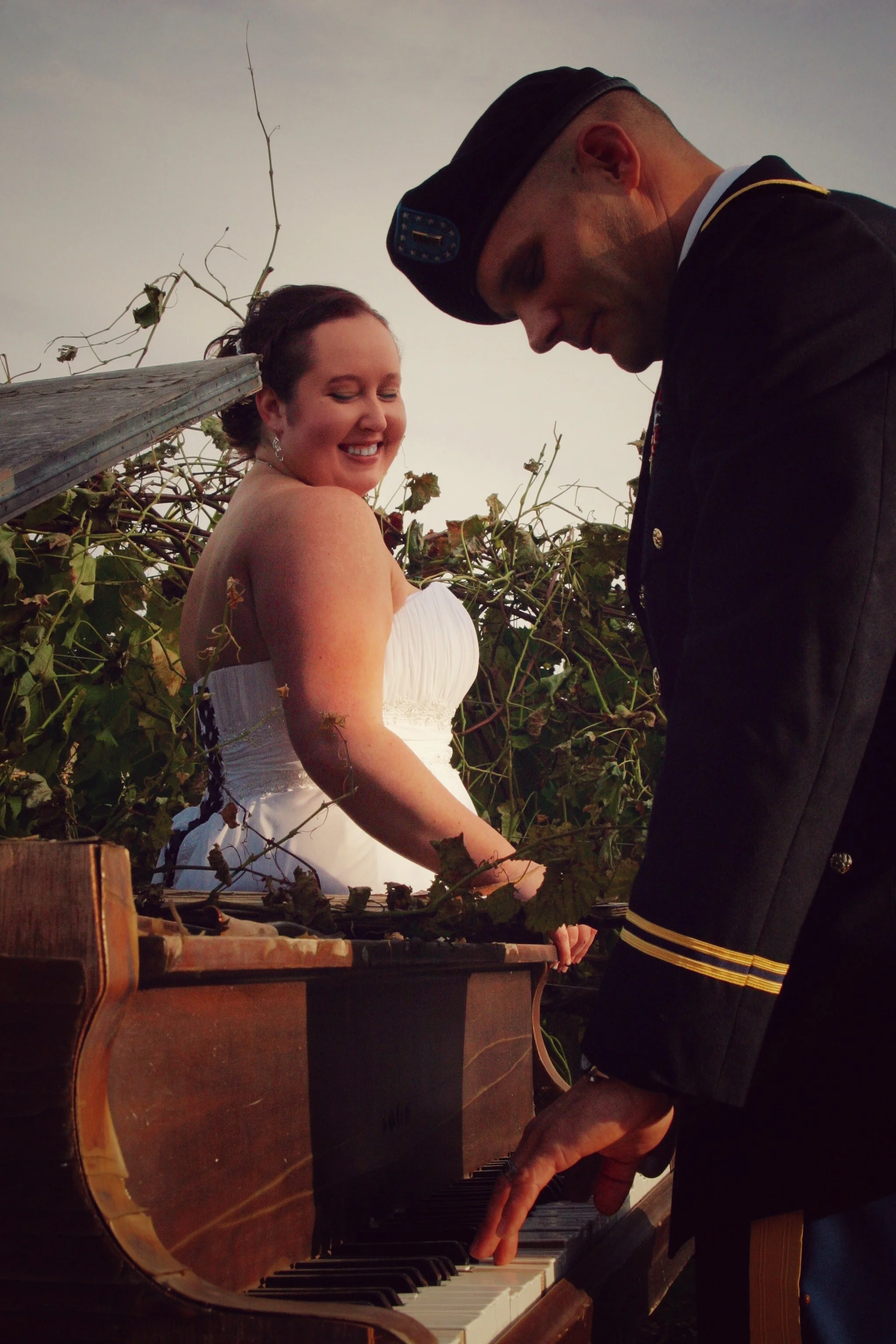 A woman in a white dress smiling while playing a piano outdoors, with a man in a military uniform playing alongside her, surrounded by vines and branches.
