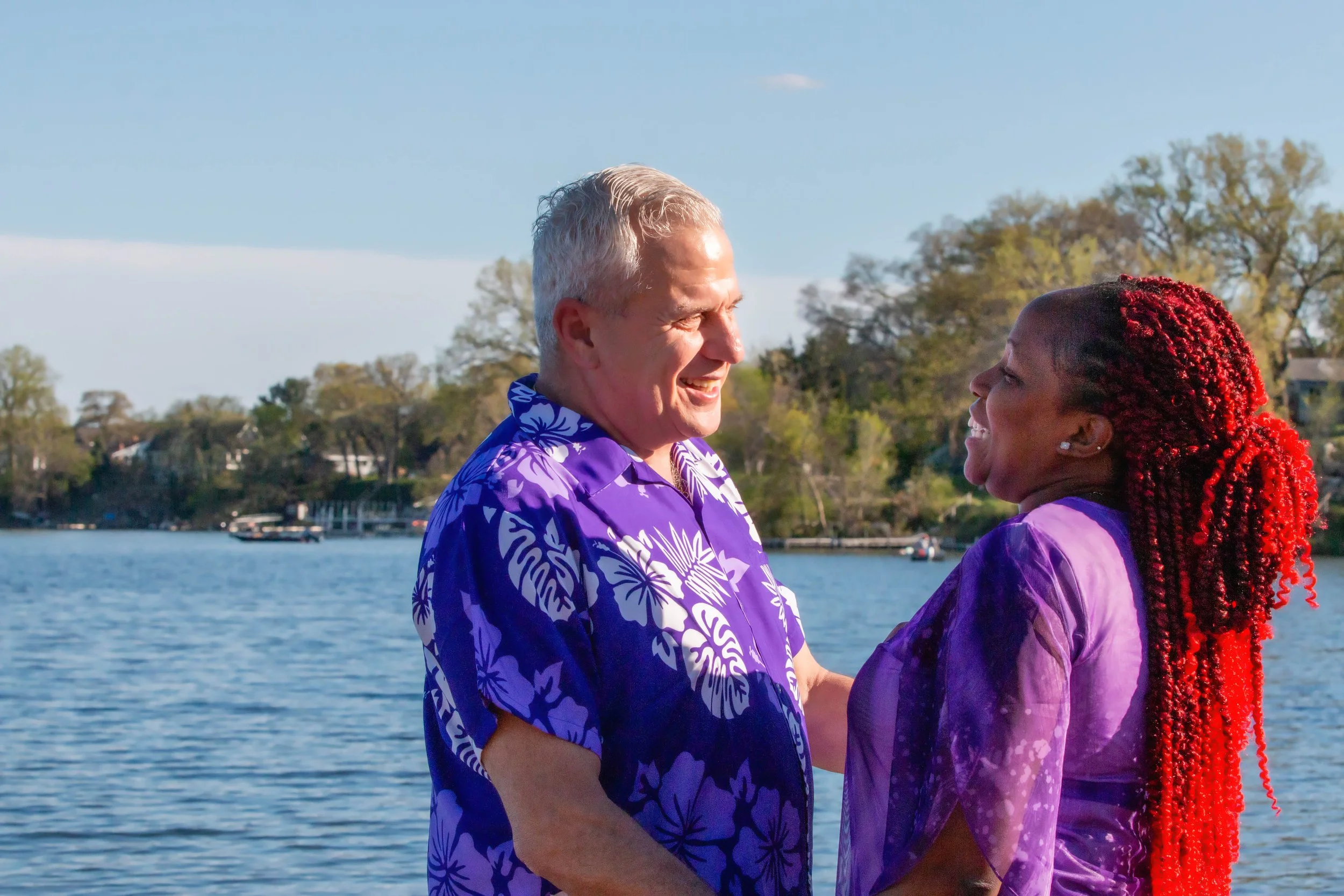 A senior man and woman sharing a joyful moment by the water, both wearing purple shirts, with trees and houses in the background.
