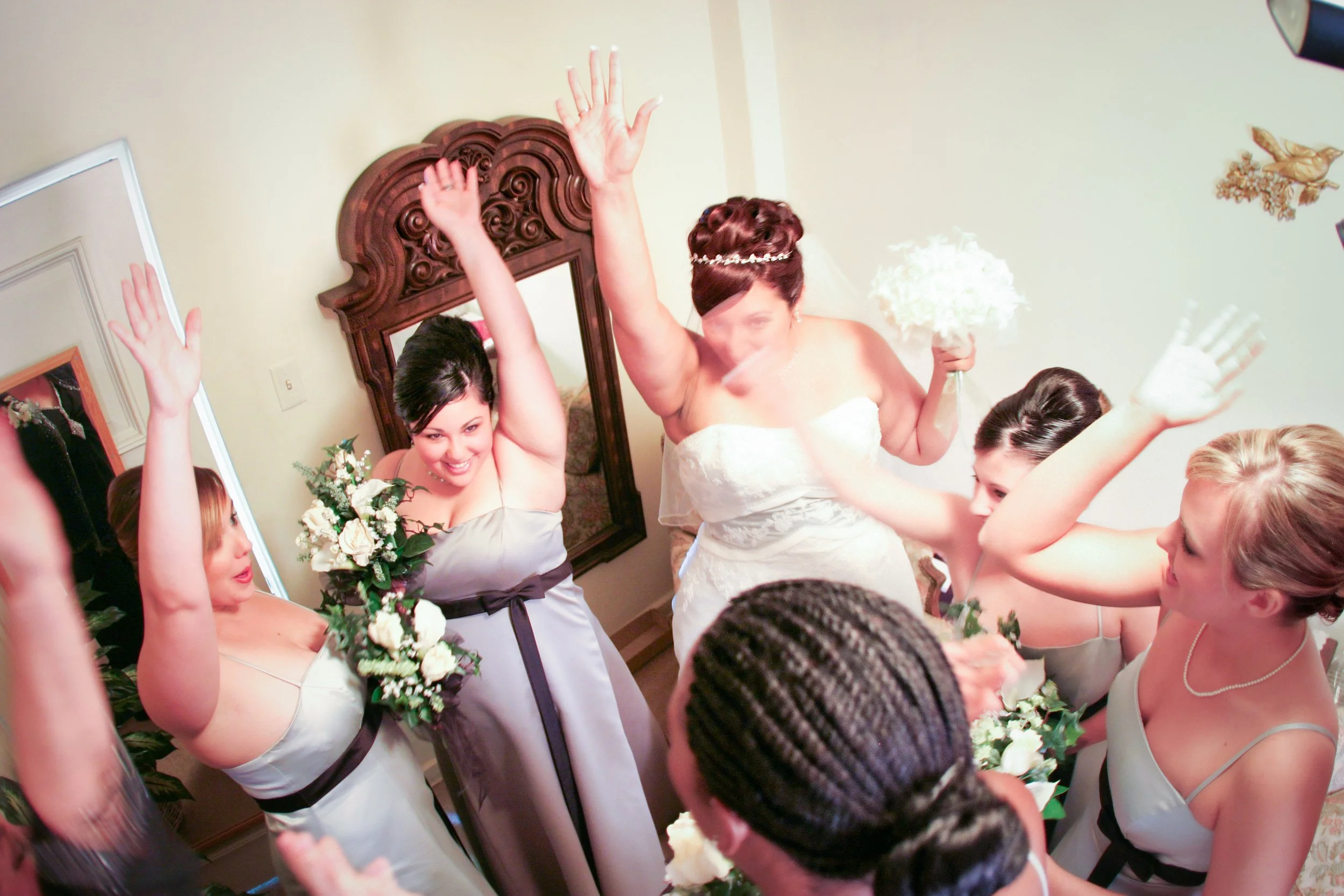 Bridal party celebrating, with women raising their hands and holding bouquets in a room decorated with a mirror and wall art.