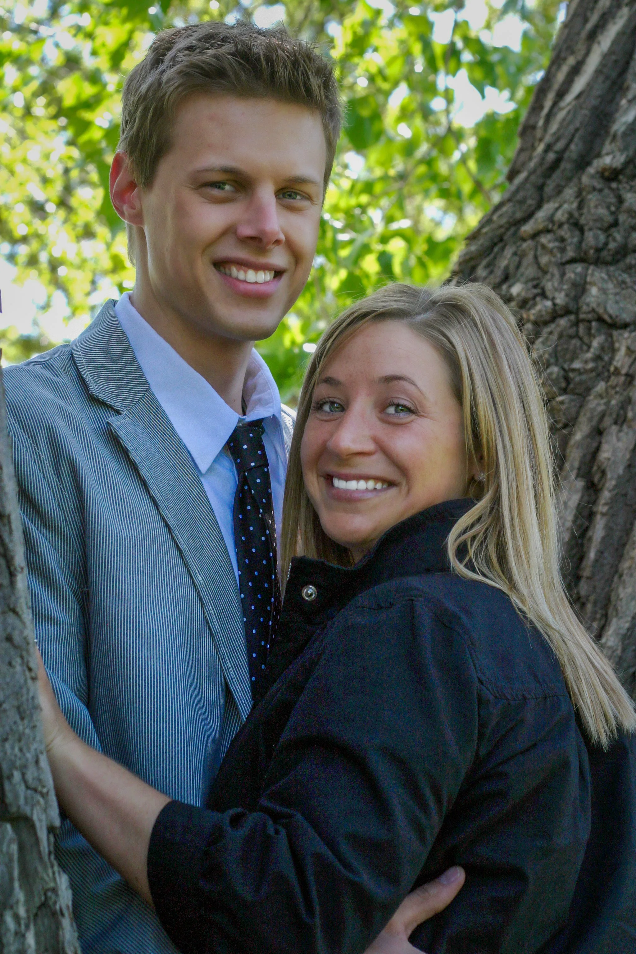 A smiling young man in a suit and tie standing next to a smiling young woman in a black jacket outdoors near a tree.
