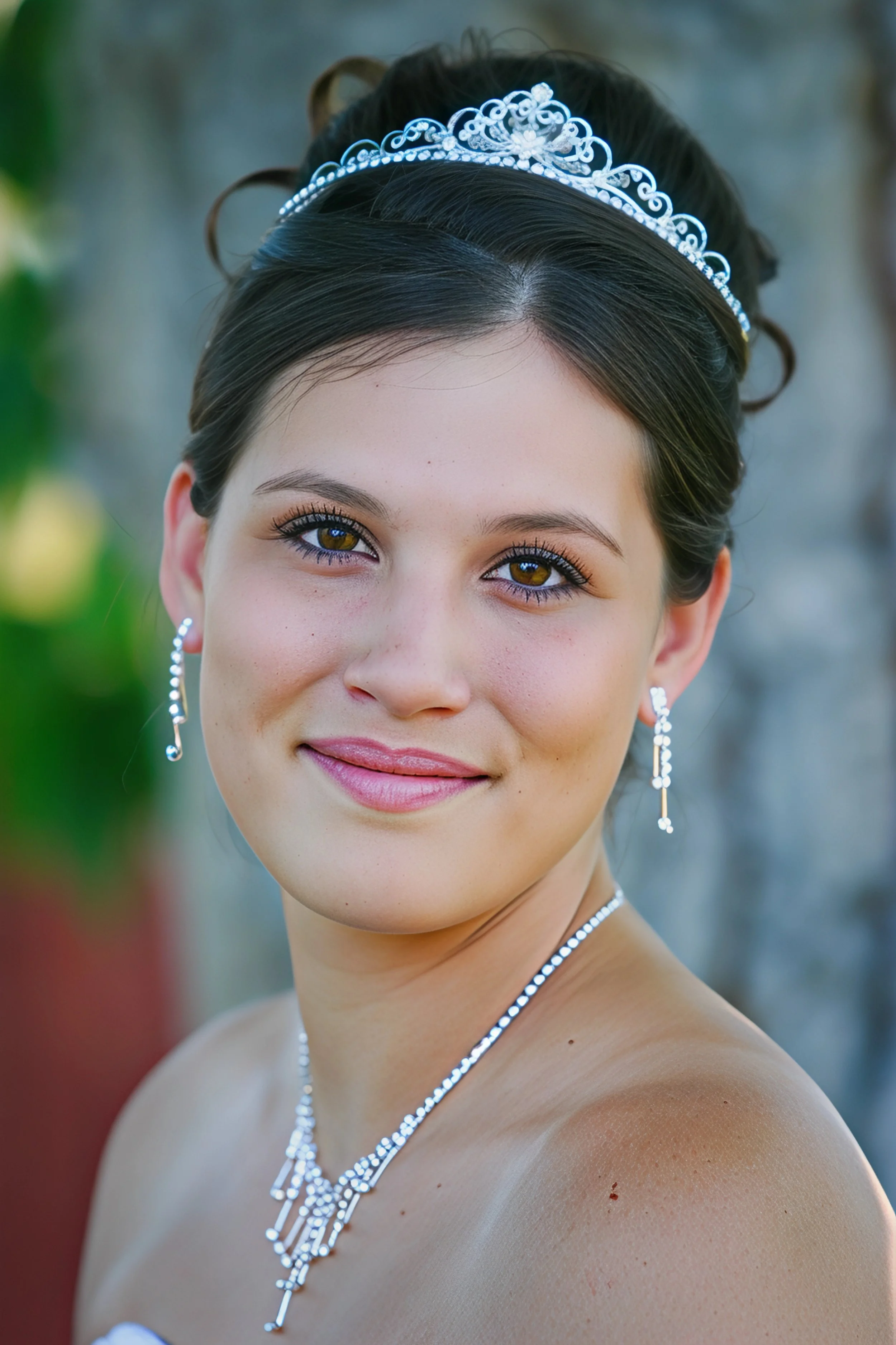 Close-up portrait of a young woman wearing a tiara, earrings, and a necklace, smiling outdoors with a blurred natural background.