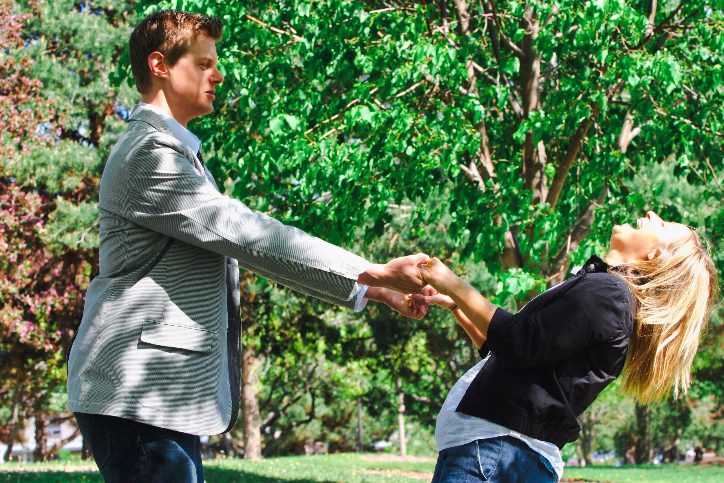 A young man in a silver blazer is holding hands with a young woman with blonde hair who is leaning back and smiling in a park with green trees and grass.