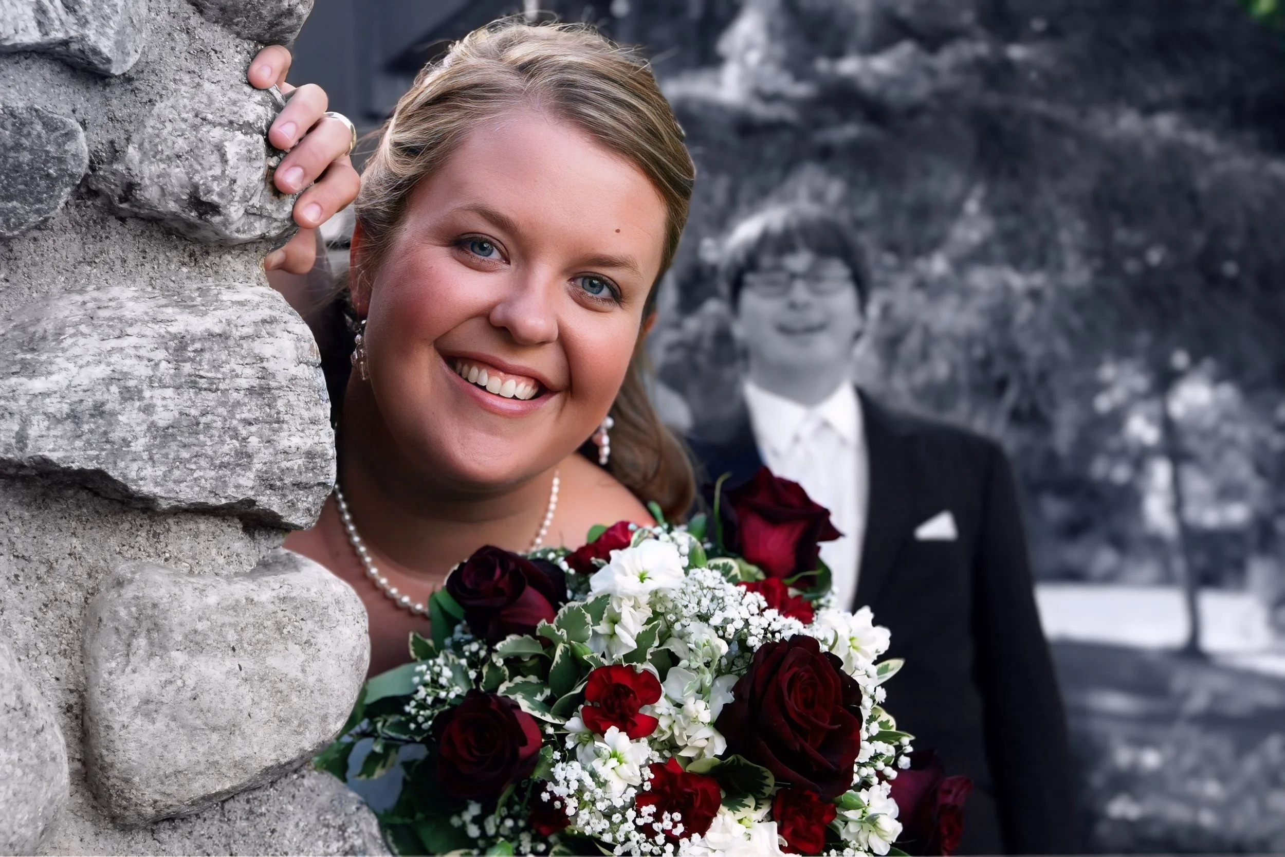 A smiling bride in a wedding dress holding a bouquet of red and white flowers, peeking from behind a stone wall, with a groom in a suit and glasses in the background.