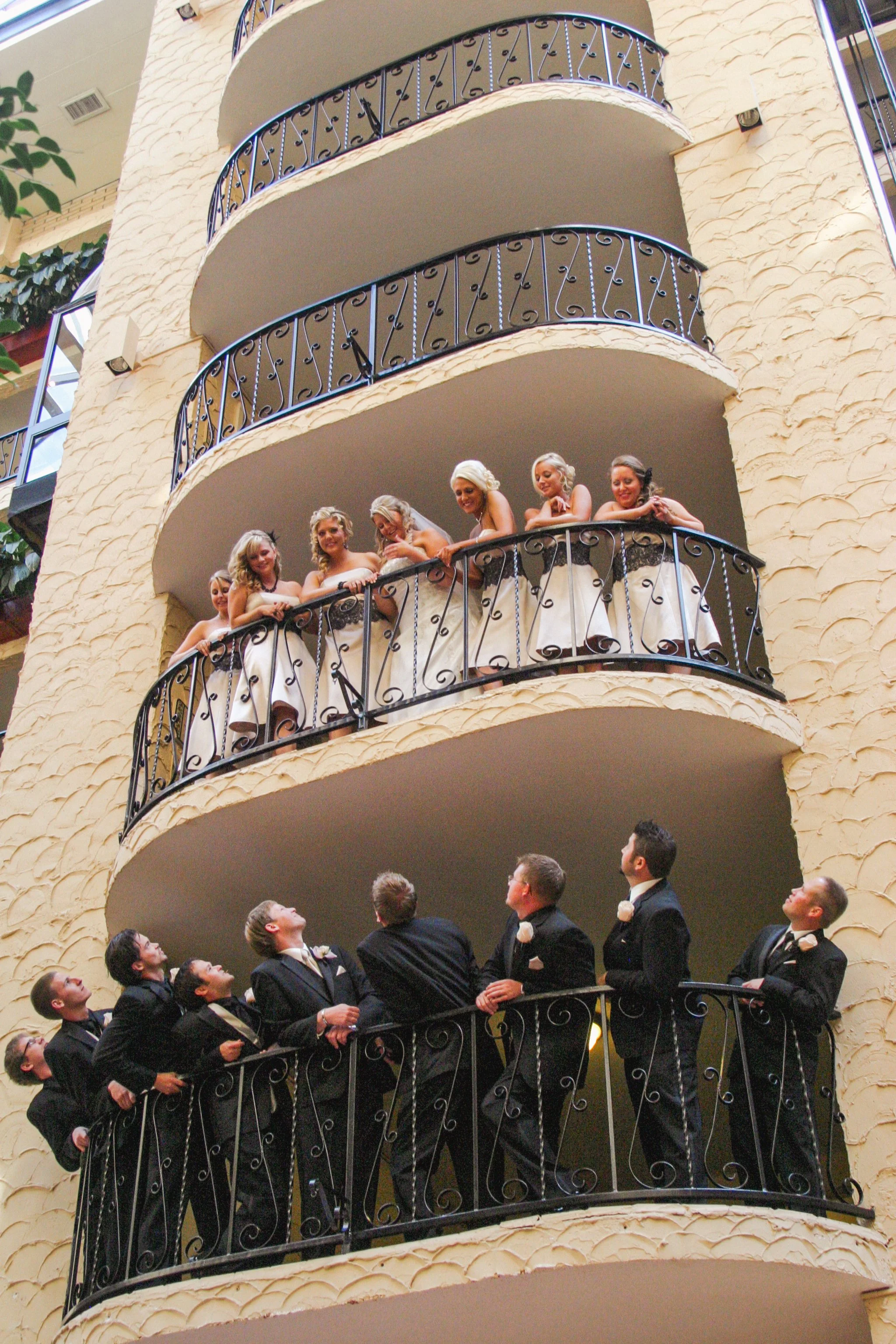 Group of bridesmaids and groomsmen on hotel balconies, dressed in formal attire, celebrating at a wedding.