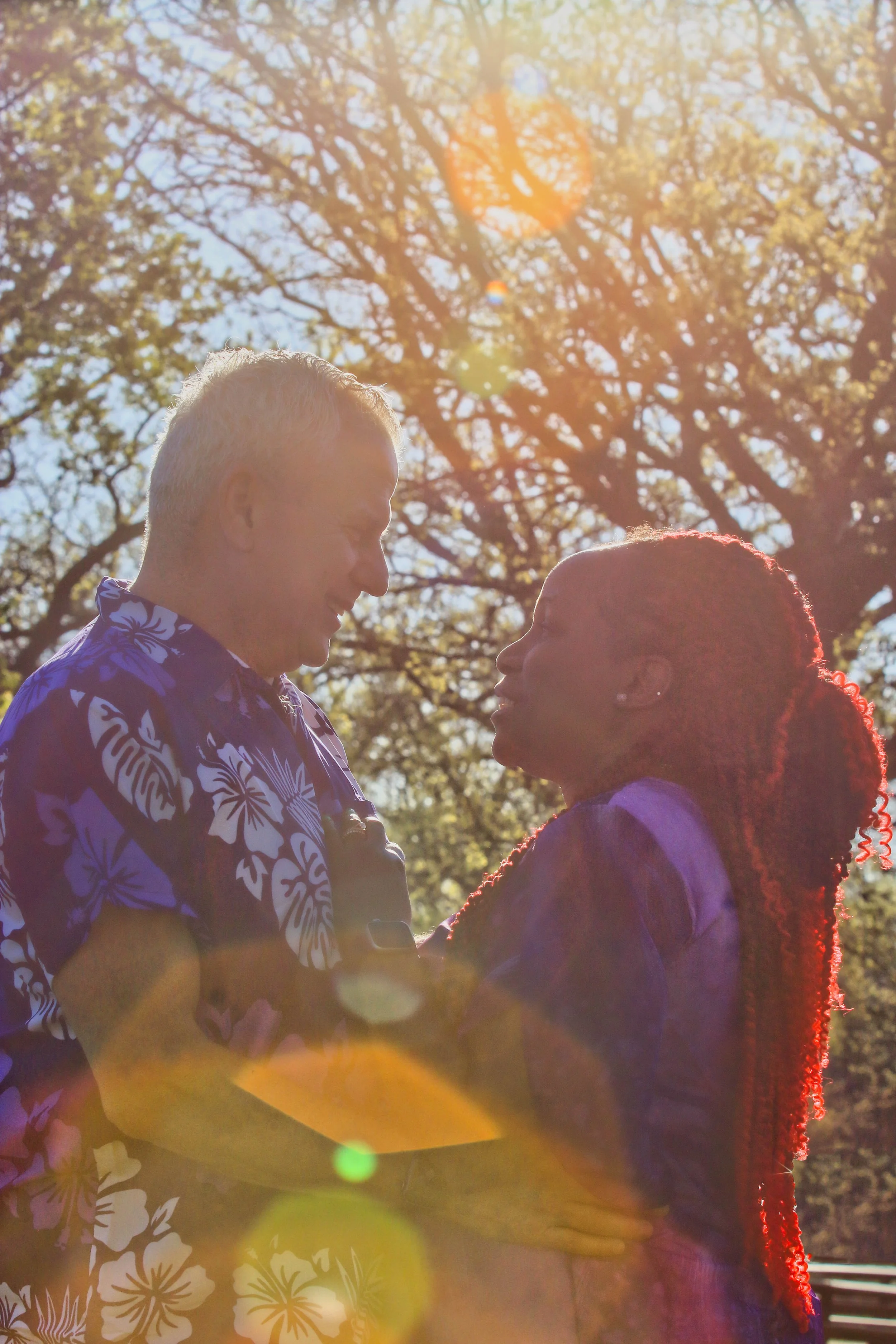 A couple sharing a moment outdoors, smiling and holding each other while sunlight filters through the trees in the background.