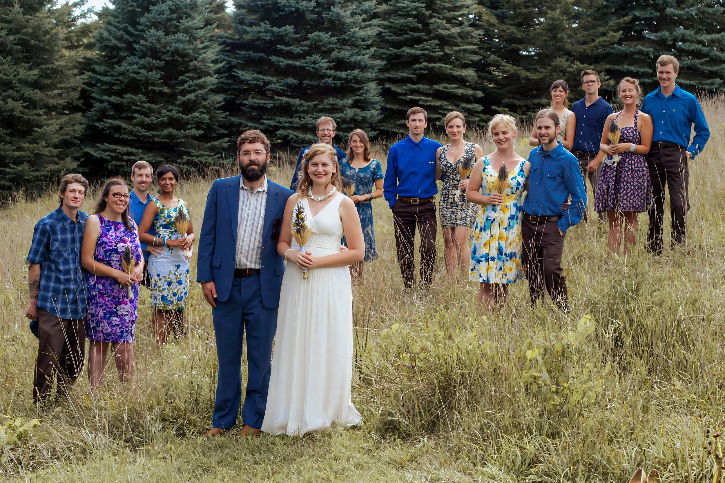 Group of people in colorful clothing standing in a grassy field with trees in the background, celebrating a wedding.