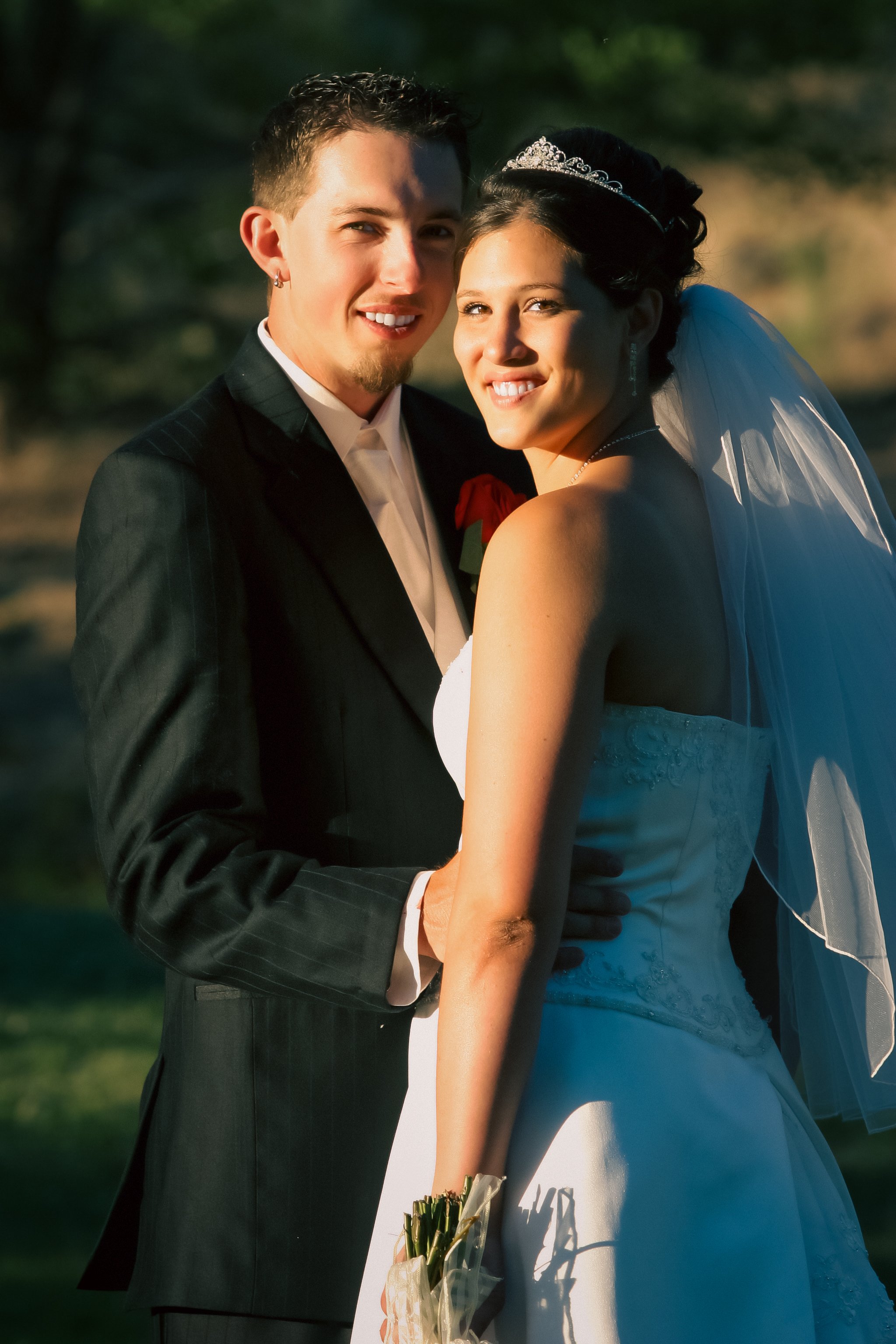 A newlywed couple stands outdoors, smiling at the camera during sunset. The groom is wearing a black suit with a white shirt, and the bride is in a white wedding gown with a veil and a tiara, holding a bouquet of flowers.