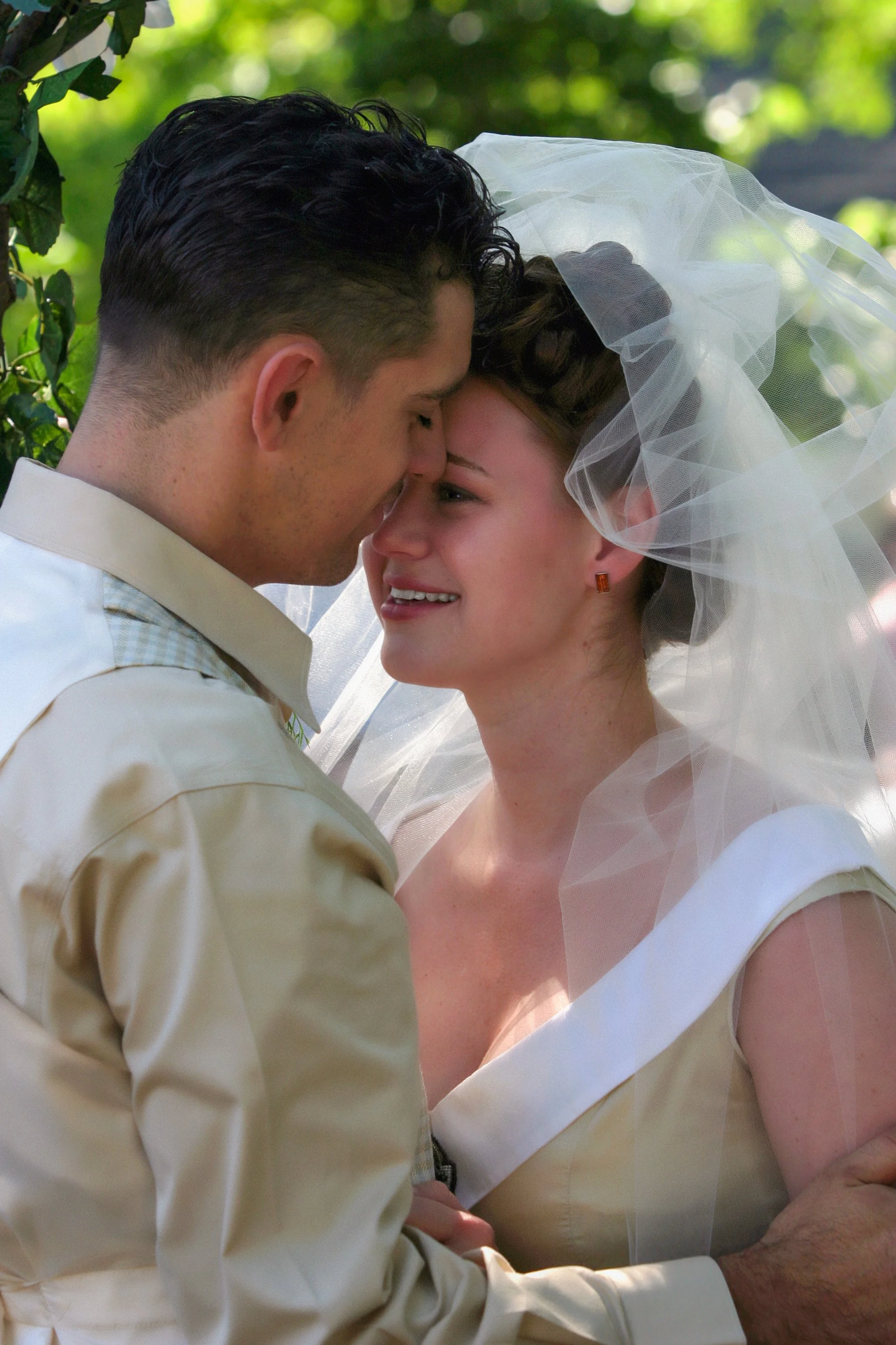 A couple on their wedding day sharing an intimate moment with foreheads touching, outdoors with green foliage in the background, the bride wearing a wedding veil and earrings, the groom in a light-colored shirt.