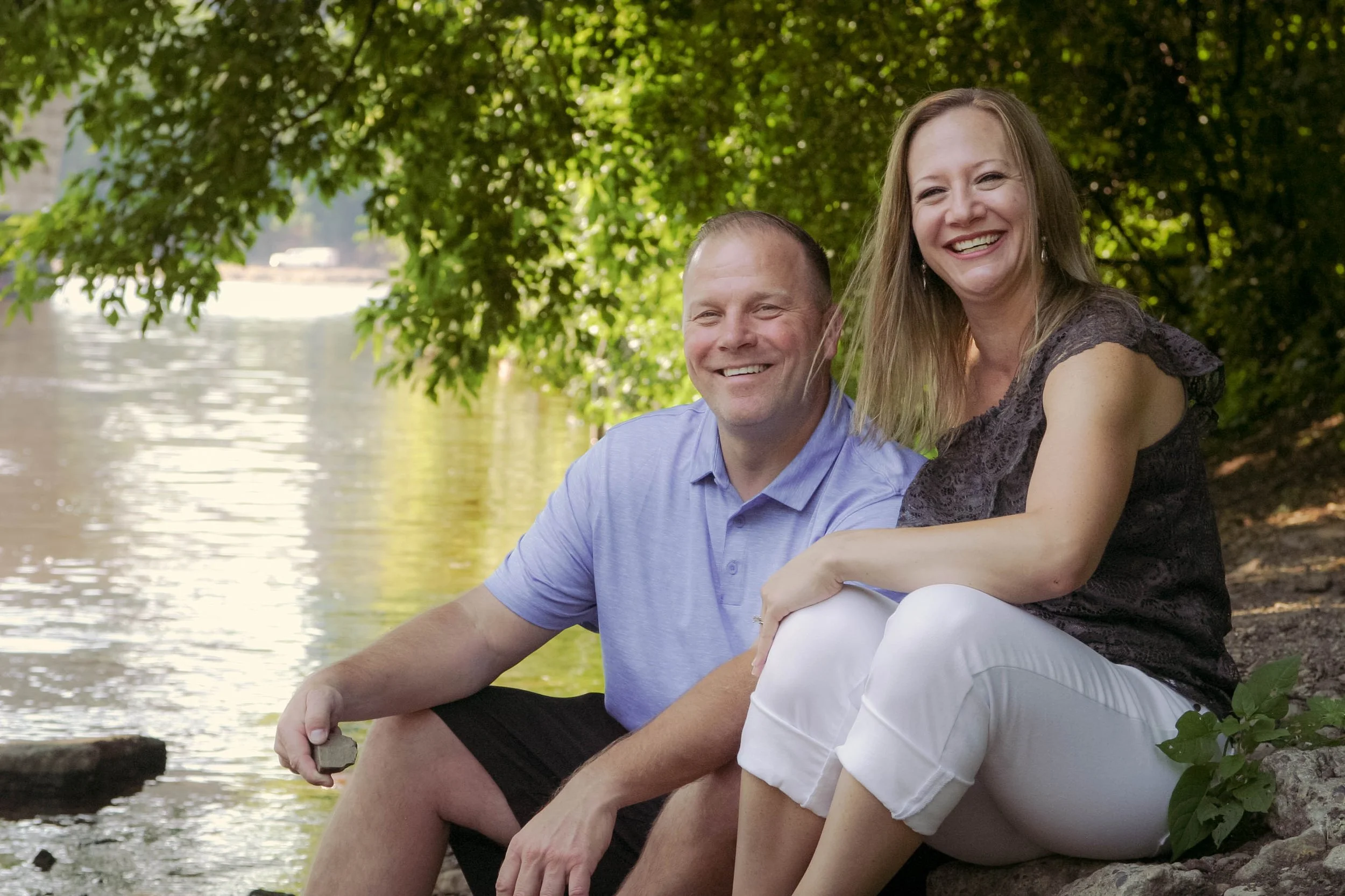 A cheerful couple sitting by a river, surrounded by trees, enjoying a sunny day.