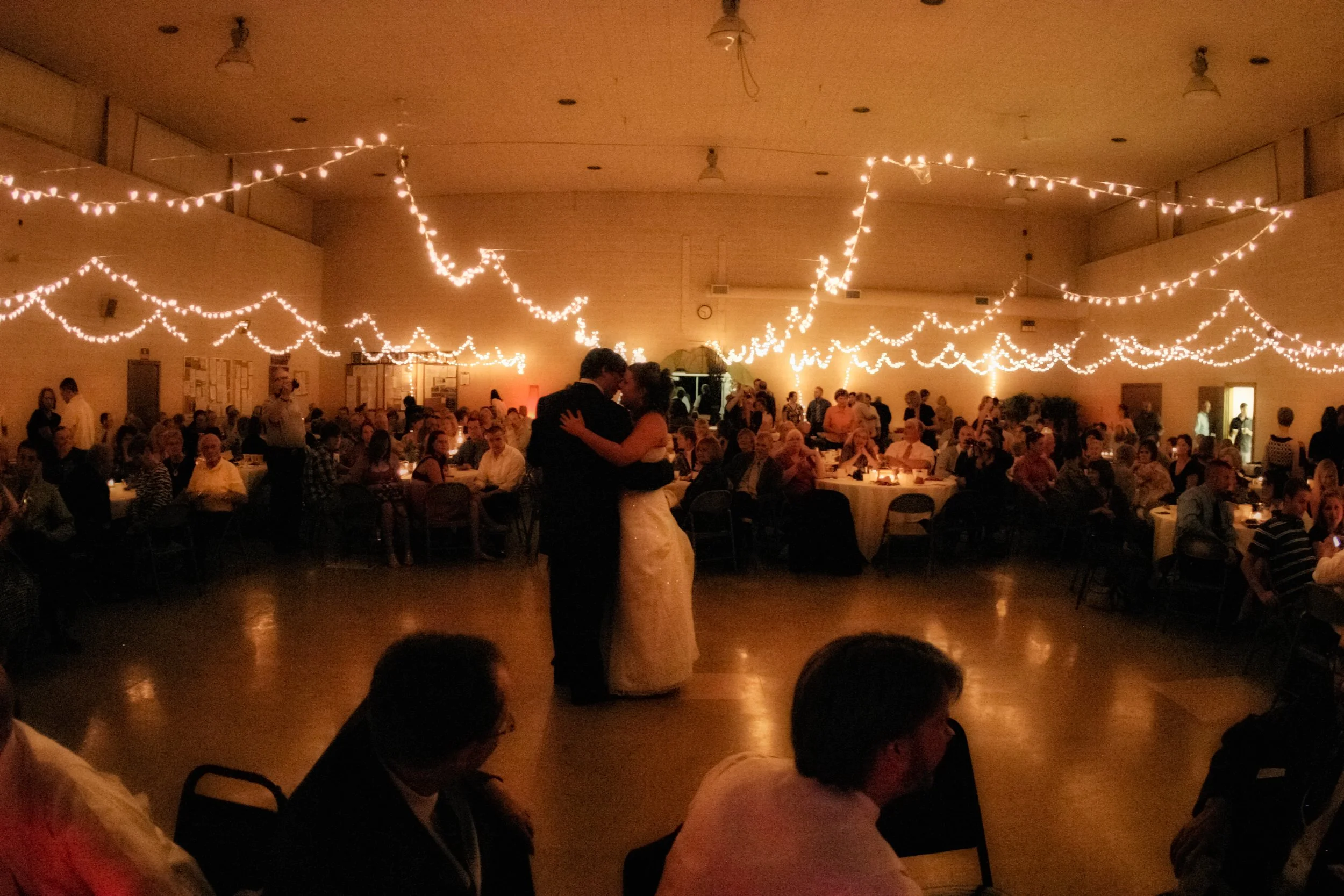 People dancing in a dimly lit hall decorated with string lights, with many seated guests observing.