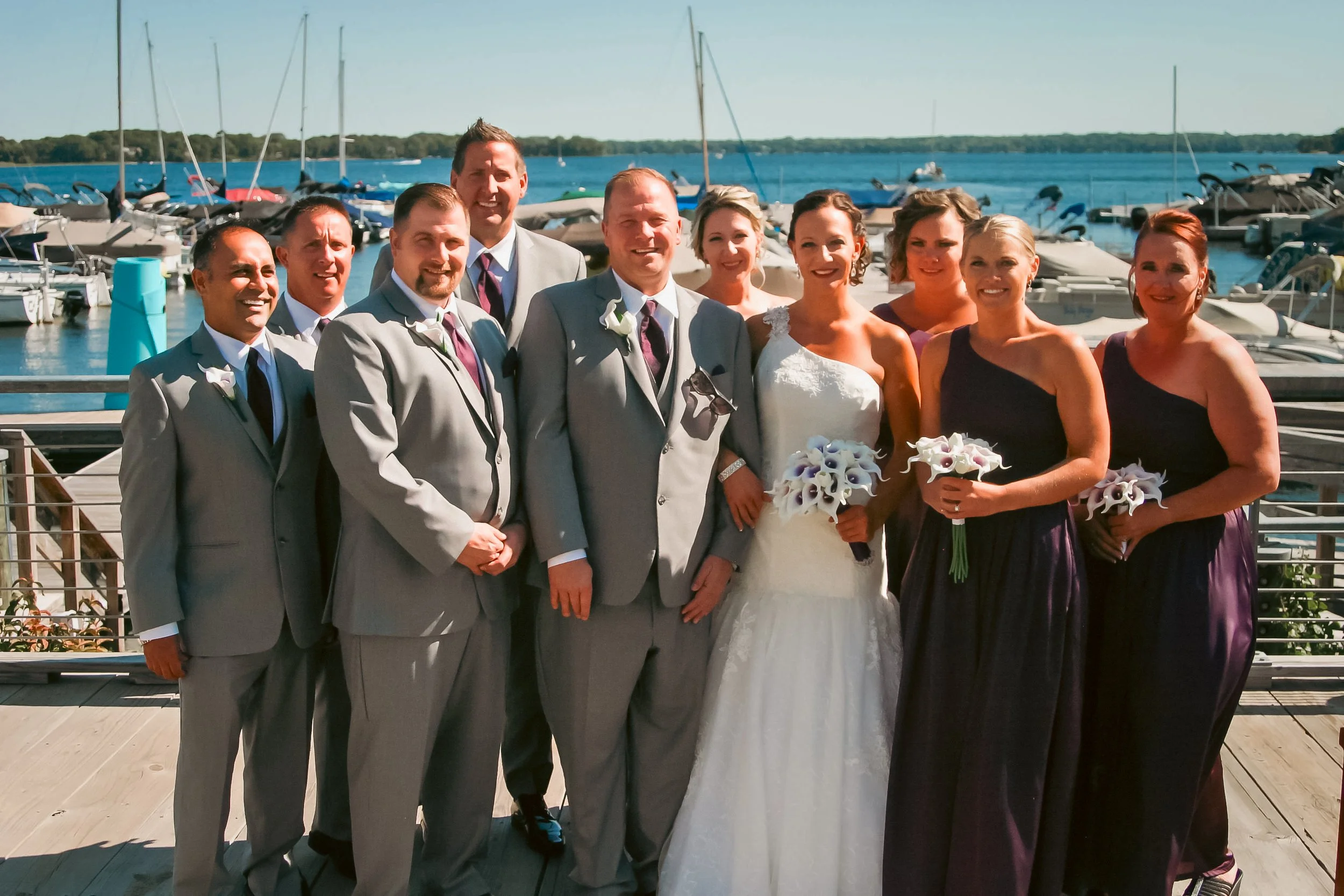 A group of people at a wedding by a marina, including the bride in a white gown holding a bouquet, the groom in a gray suit, and bridesmaids in dark dresses holding bouquets, with sailboats and the water in the background.
