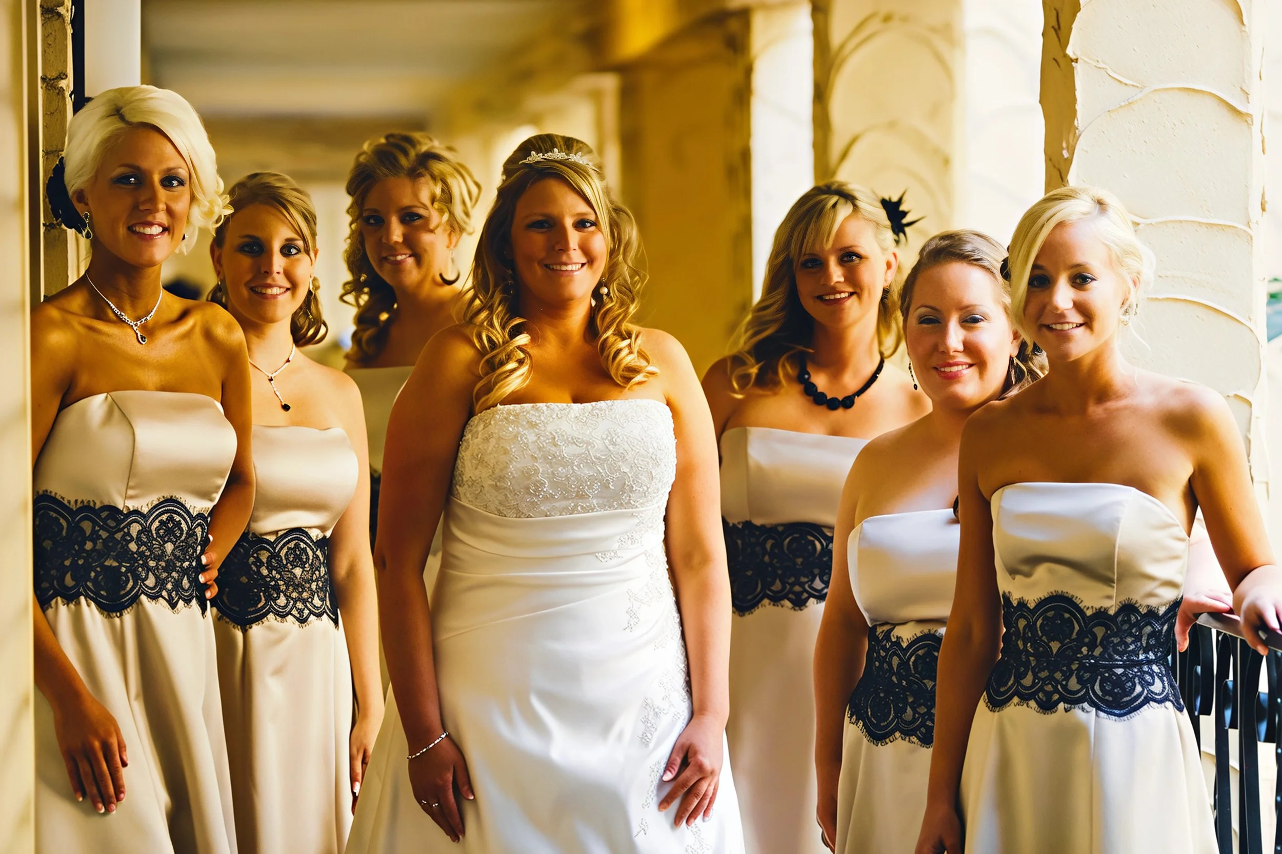 A bride in a white wedding gown standing with her bridesmaids in matching strapless cream dresses with black lace accents, inside a building with stone columns.