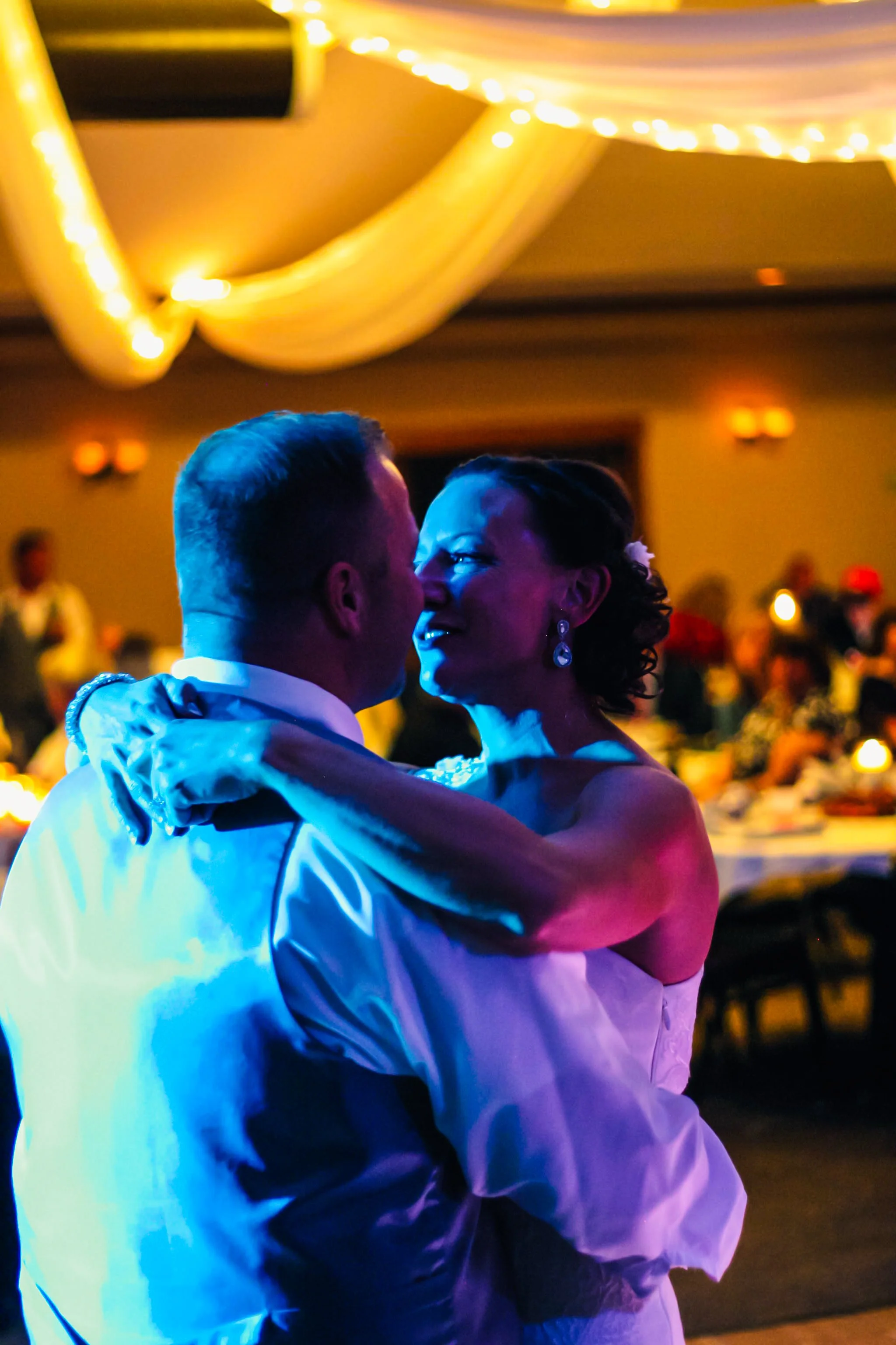 A bride and groom are dancing closely at their wedding reception, illuminated by colorful lights with draped fabric and string lights overhead in the background.