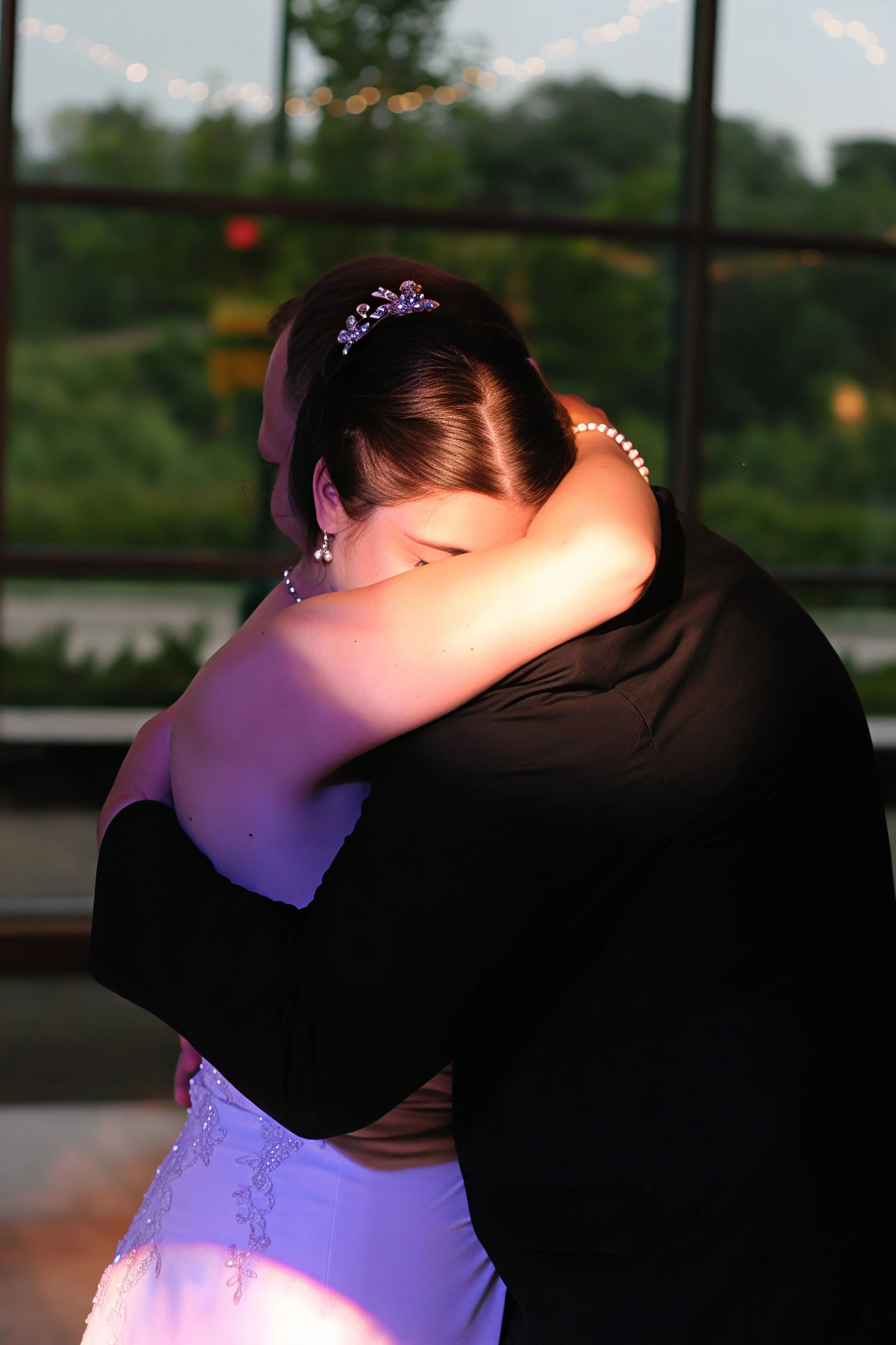 A bride and groom hug closely during their wedding, with the bride wearing a white dress and the groom in a black suit, inside a venue with large windows showing a green outdoor area.