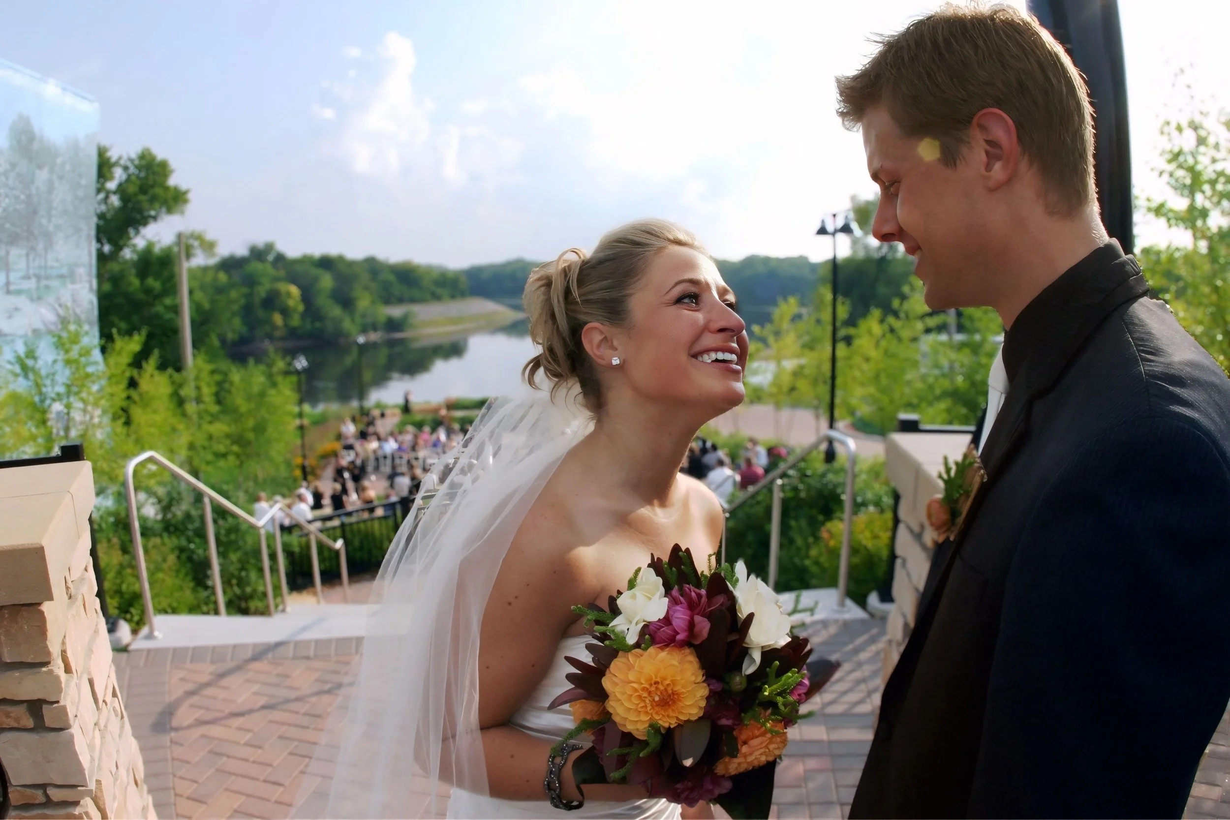 A bride and groom sharing a moment outdoors during their wedding, with the bride holding a bouquet and smiling at the groom near a river and a crowd of guests in the background.