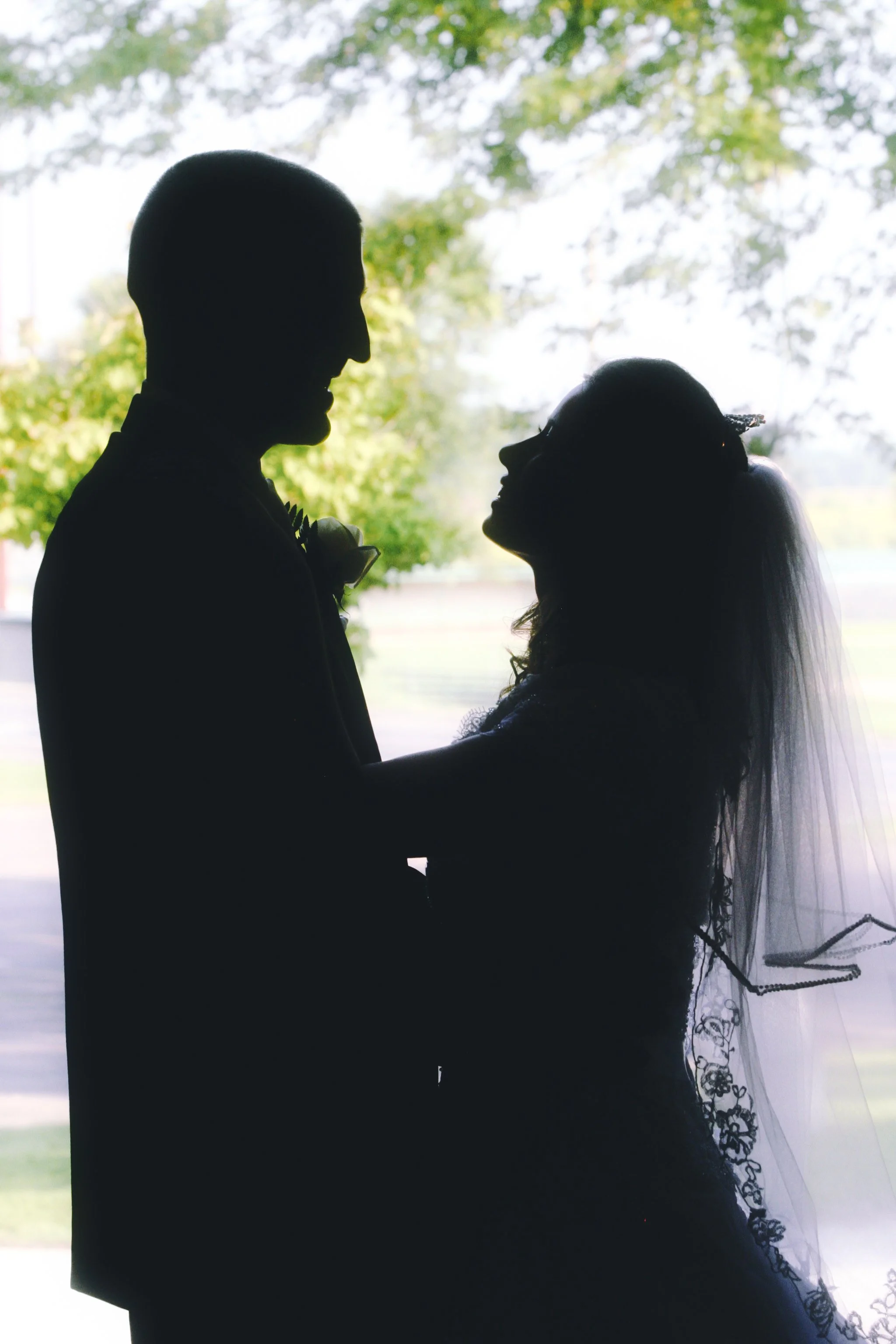 Silhouette of a bride and groom on their wedding day, standing close and looking at each other, with a bright outdoor background.