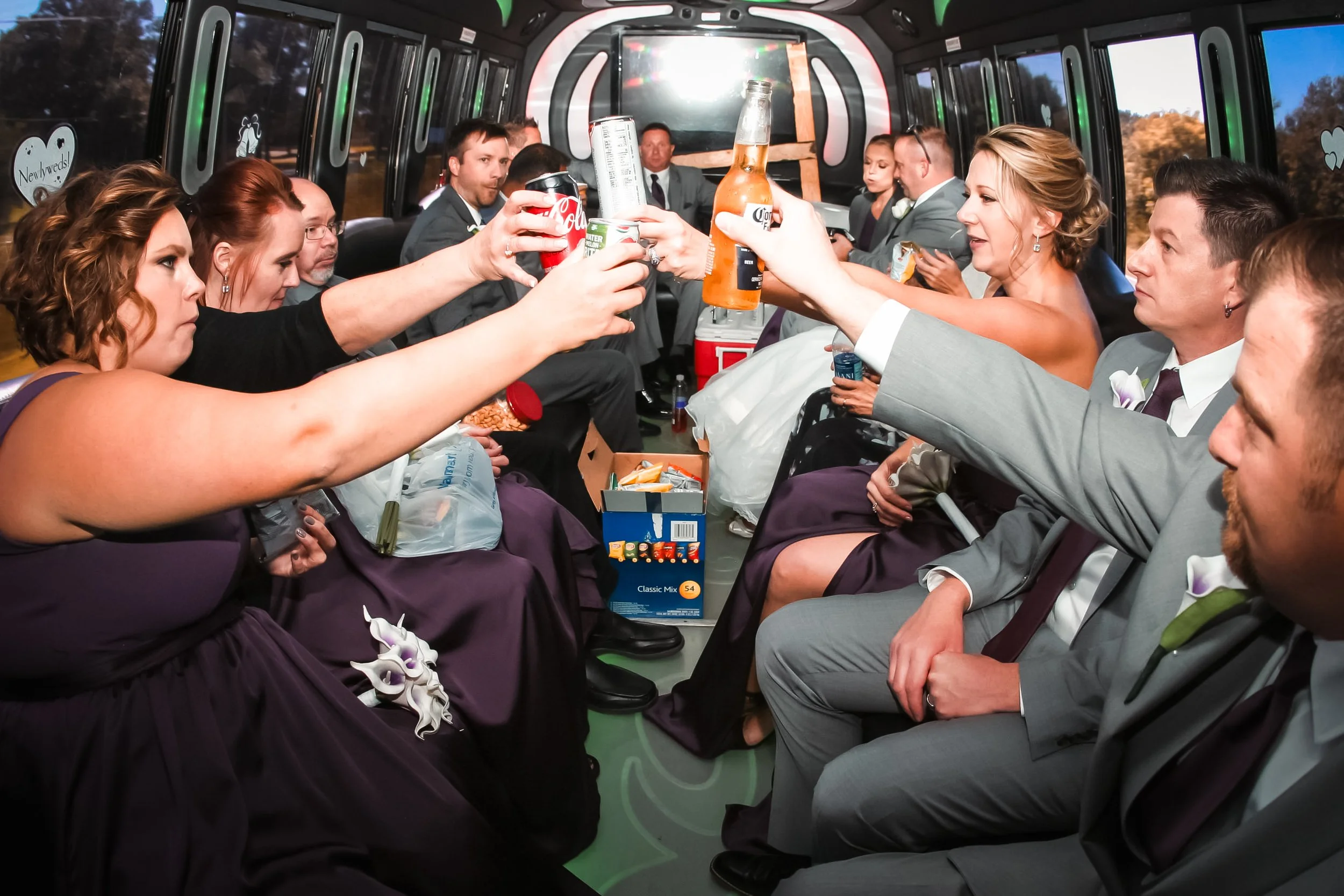 A group of wedding guests sitting inside a party bus, making a toast with drinks, some holding beer bottles and cans. They are dressed in formal attire, with women in purple dresses and men in gray suits. The bus is decorated for a celebration, likel
