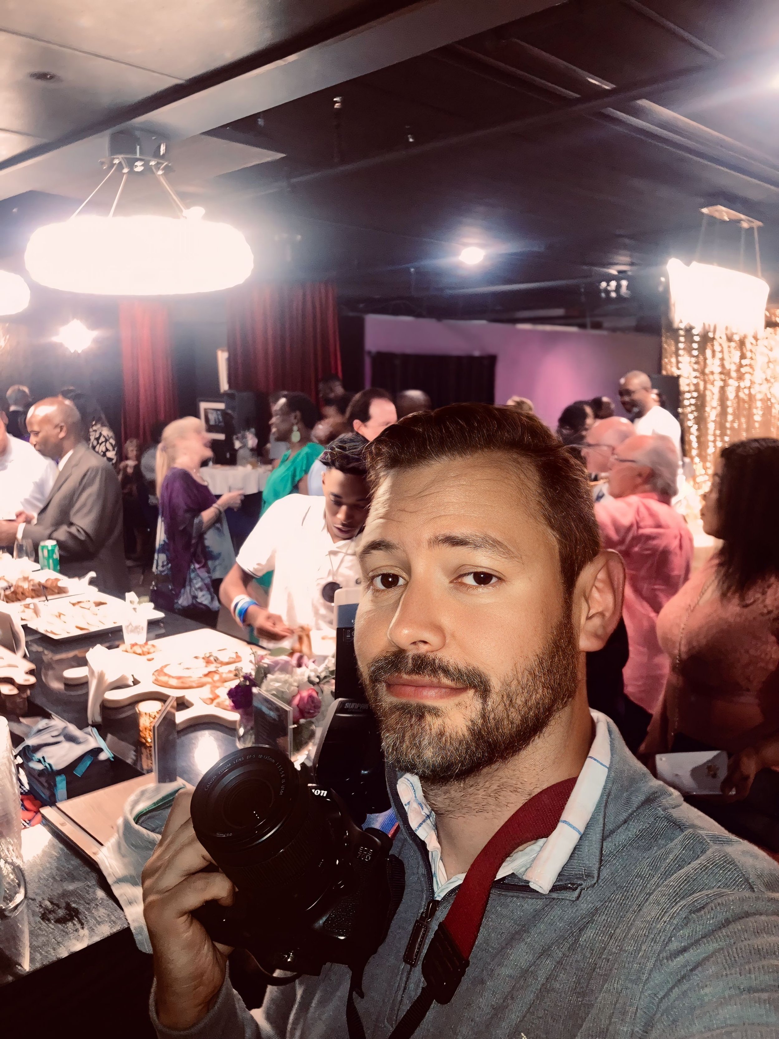 A man with a beard taking a selfie at a crowded indoor event with people gathered around tables, food, and decorations in the background.
