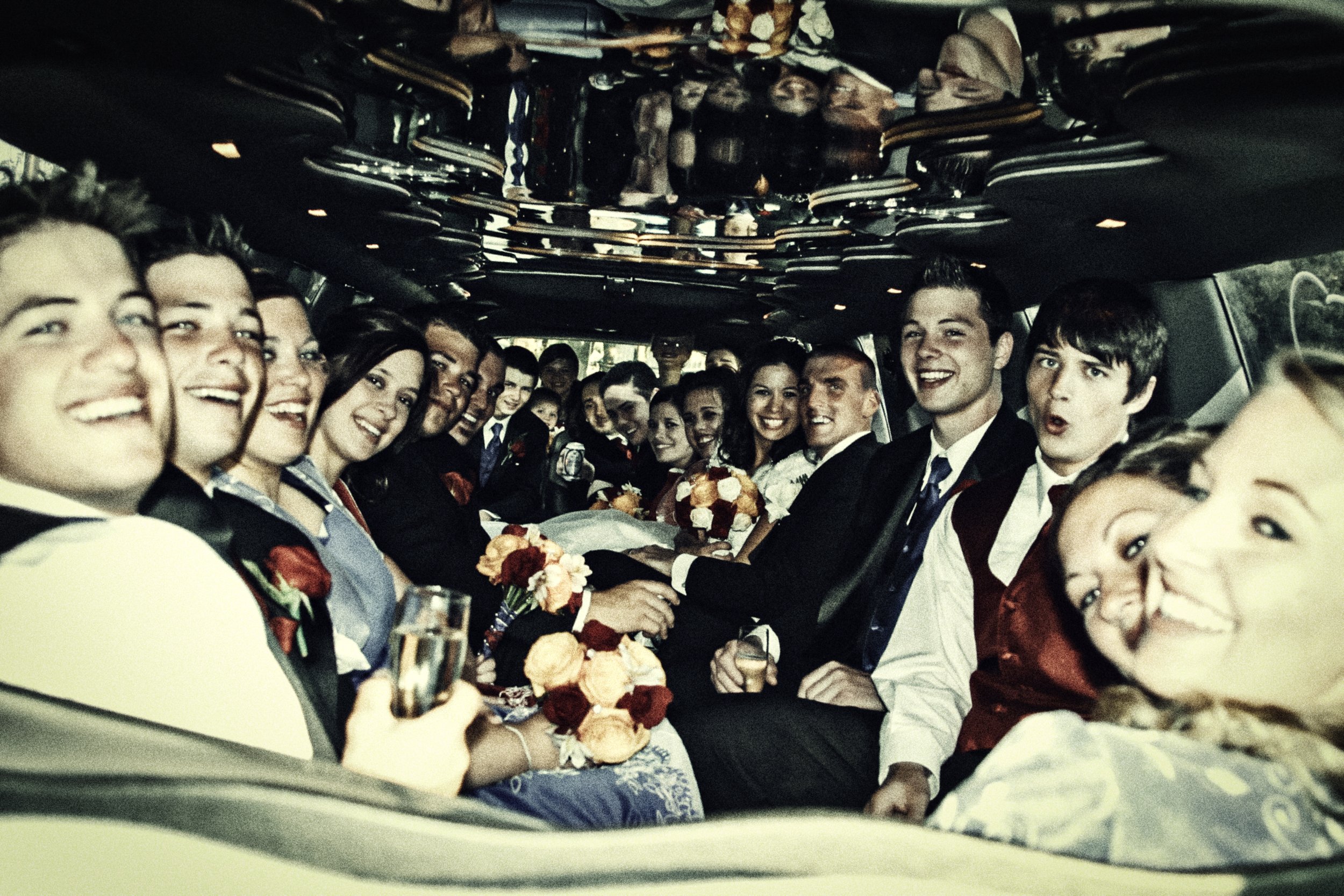 Group of people dressed in formal attire, sitting inside a luxury vehicle, smiling and holding flowers and drinks, likely celebrating a wedding.