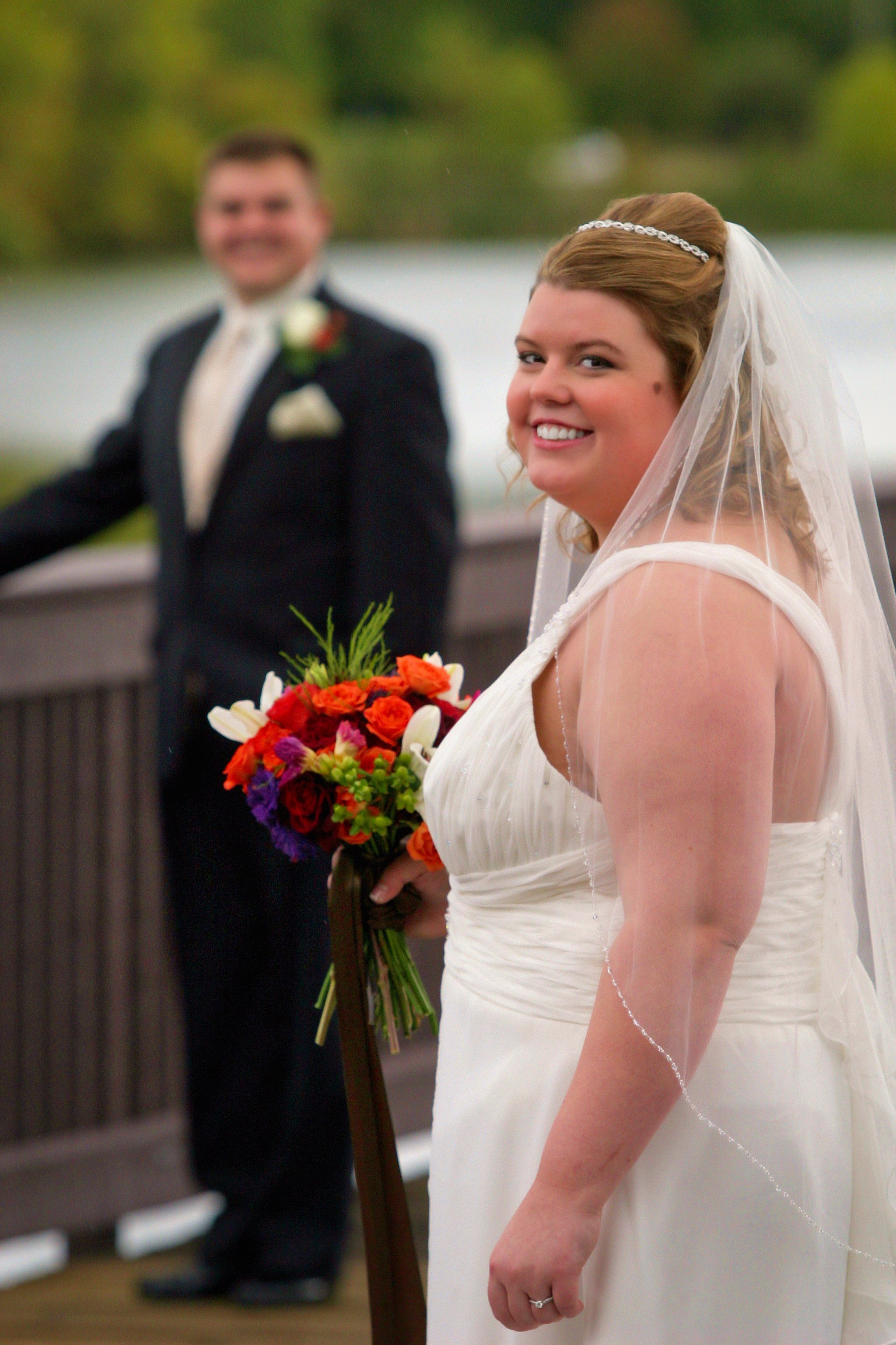 A bride in a white wedding dress holding a colorful bouquet of flowers, smiling at the camera, with a groom in a black tuxedo blurred in the background, outdoors near a lake.