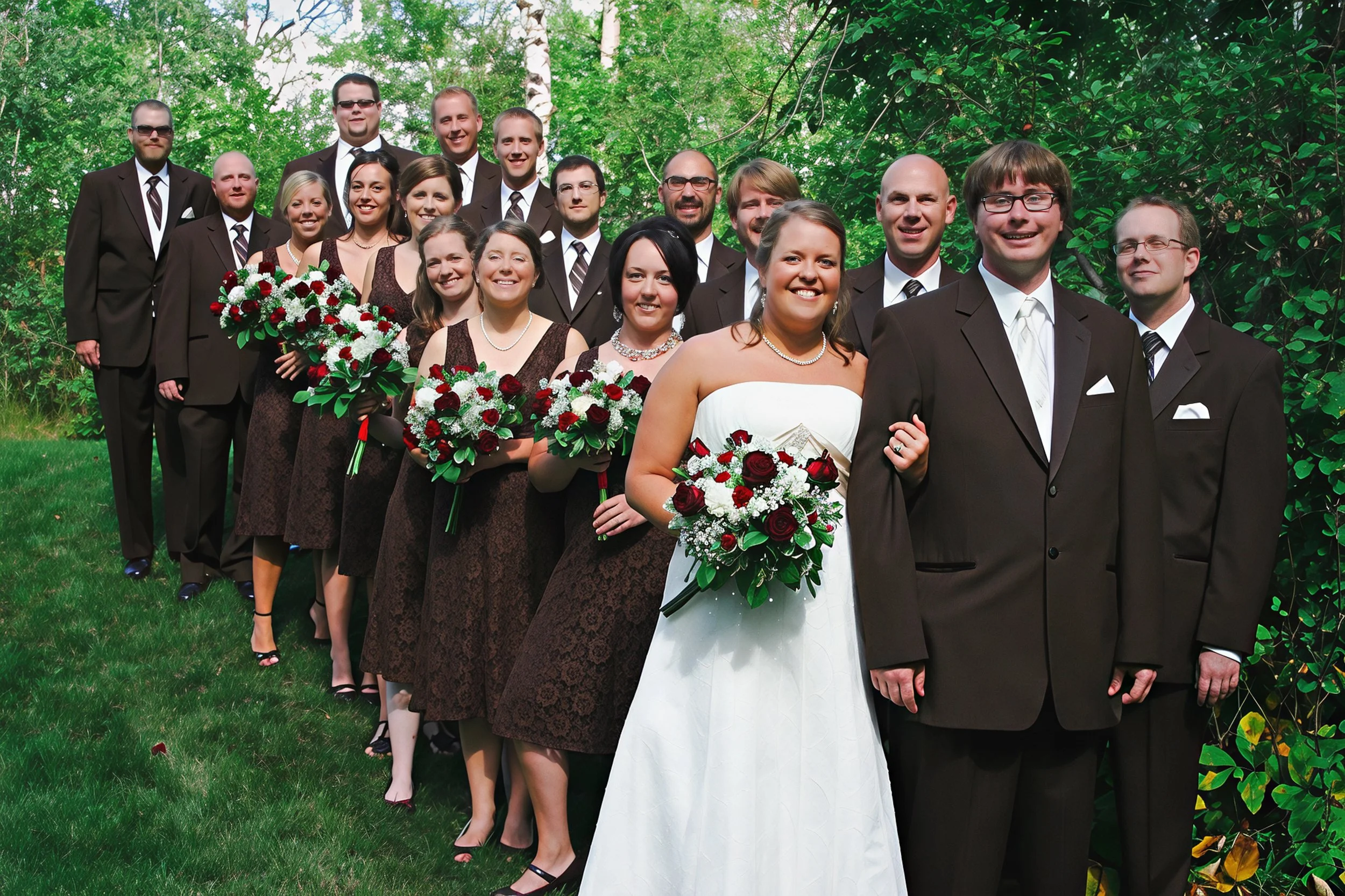 A wedding party of 16 people, including the bride in a white gown holding a bouquet, and the groom in a dark suit, standing outdoors on grass with green trees in the background. The women are holding bouquets, and all members are dressed in formal at