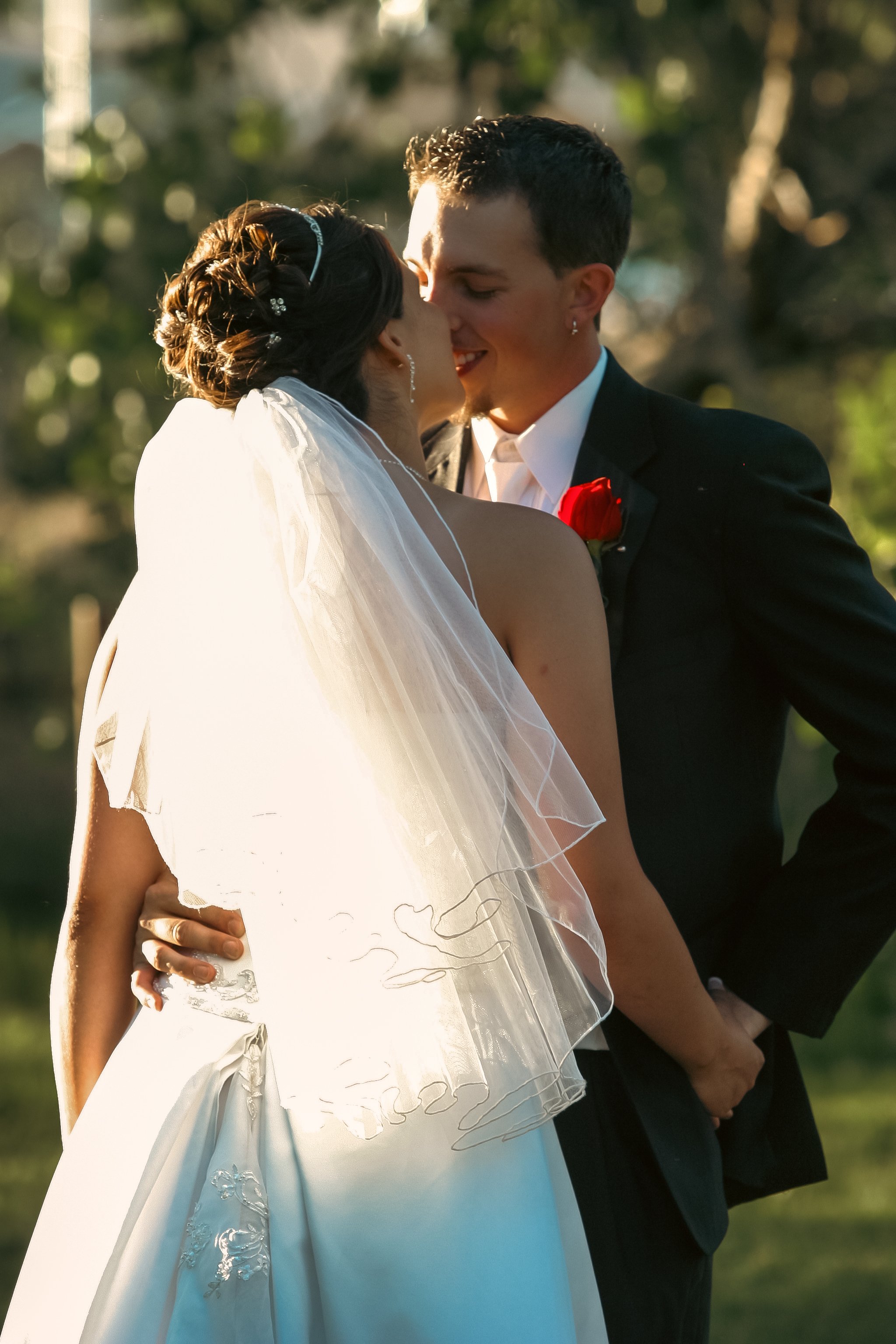 A bride and groom sharing a kiss outdoors during their wedding, with sunlight illuminating the scene and trees in the background.