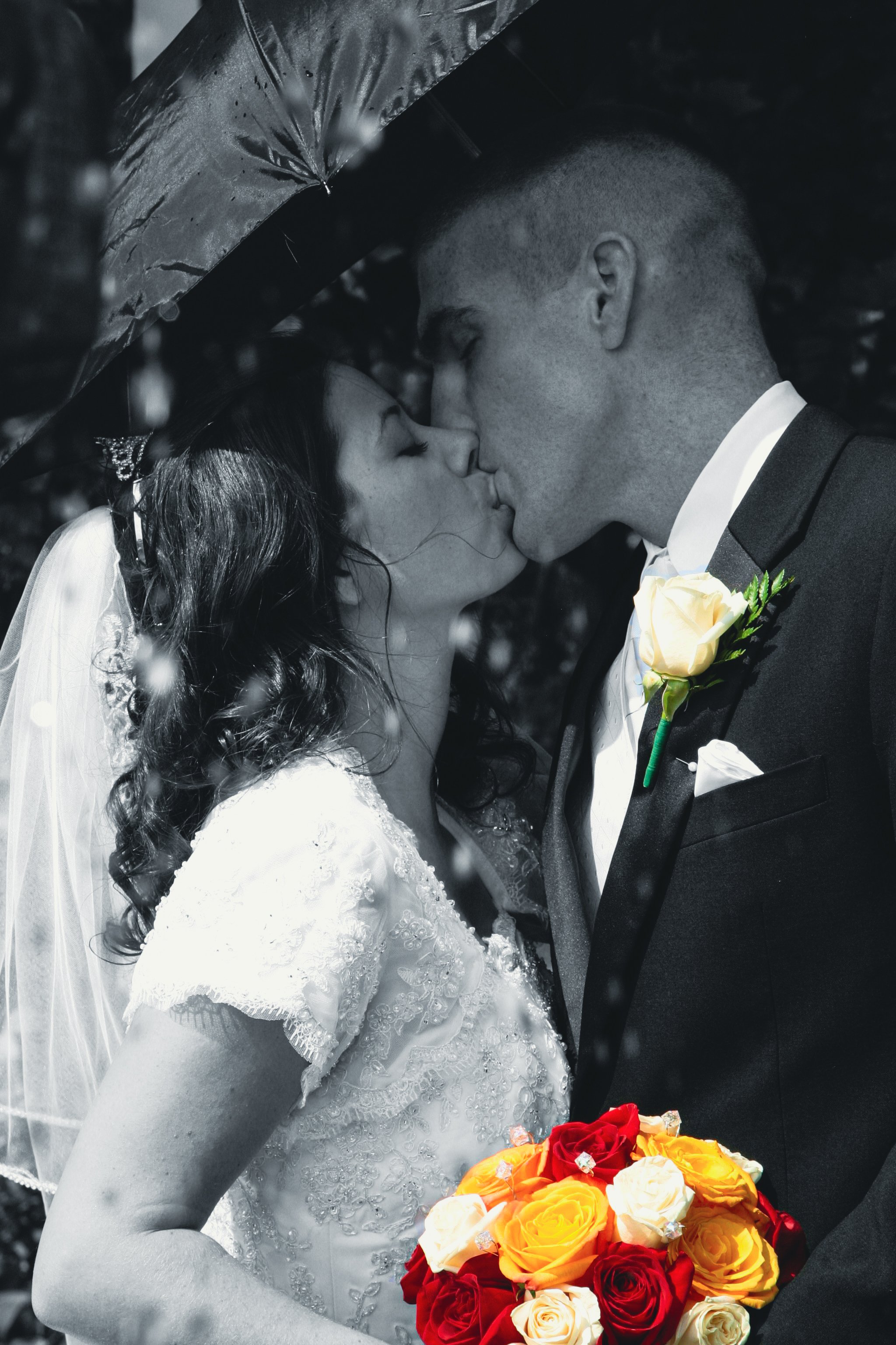 Black and white photo of a bride and groom sharing a kiss under an umbrella, with the bride holding a colorful bouquet of roses.