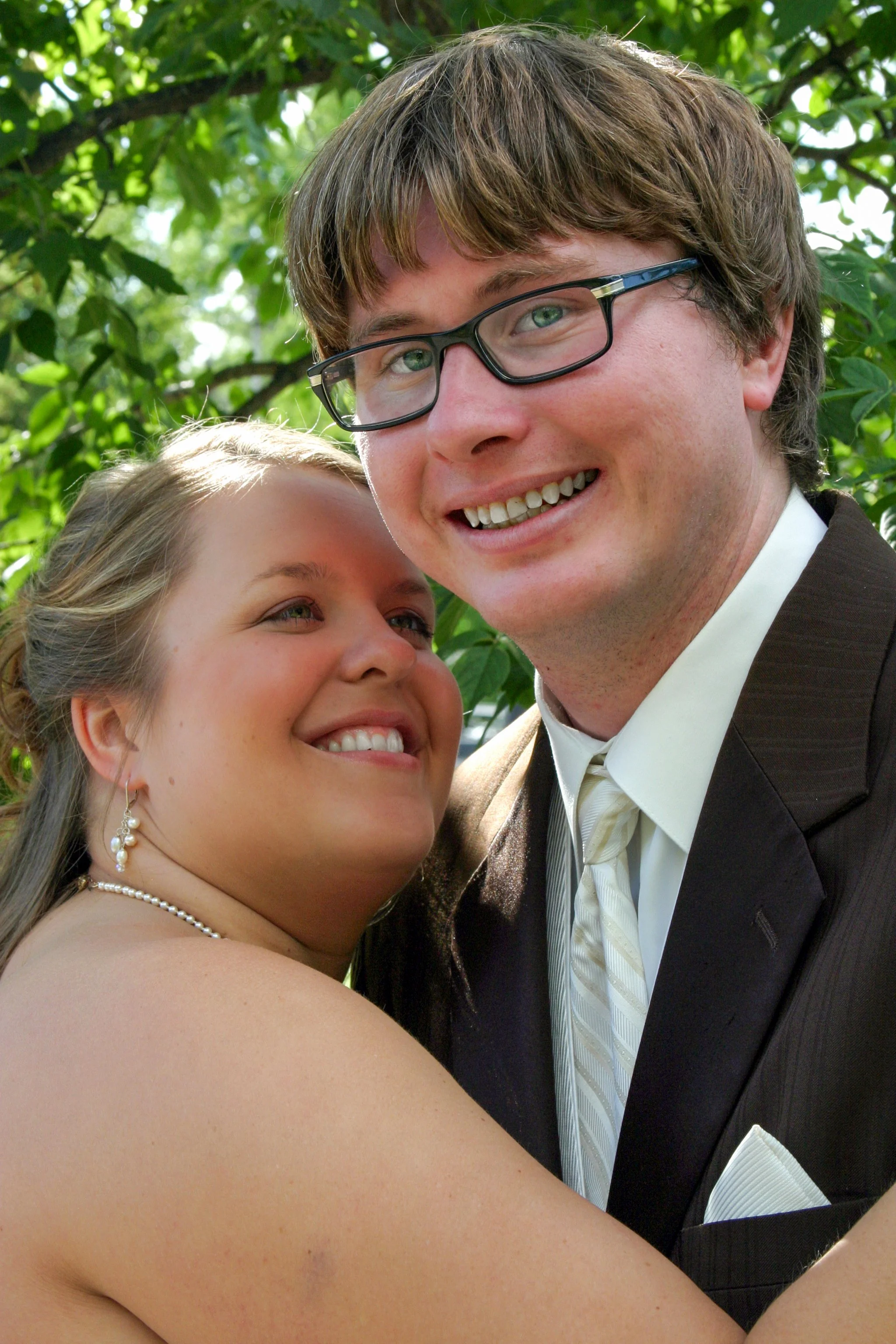 A happy bride and groom smiling and hugging outdoors, with green leaves in the background.