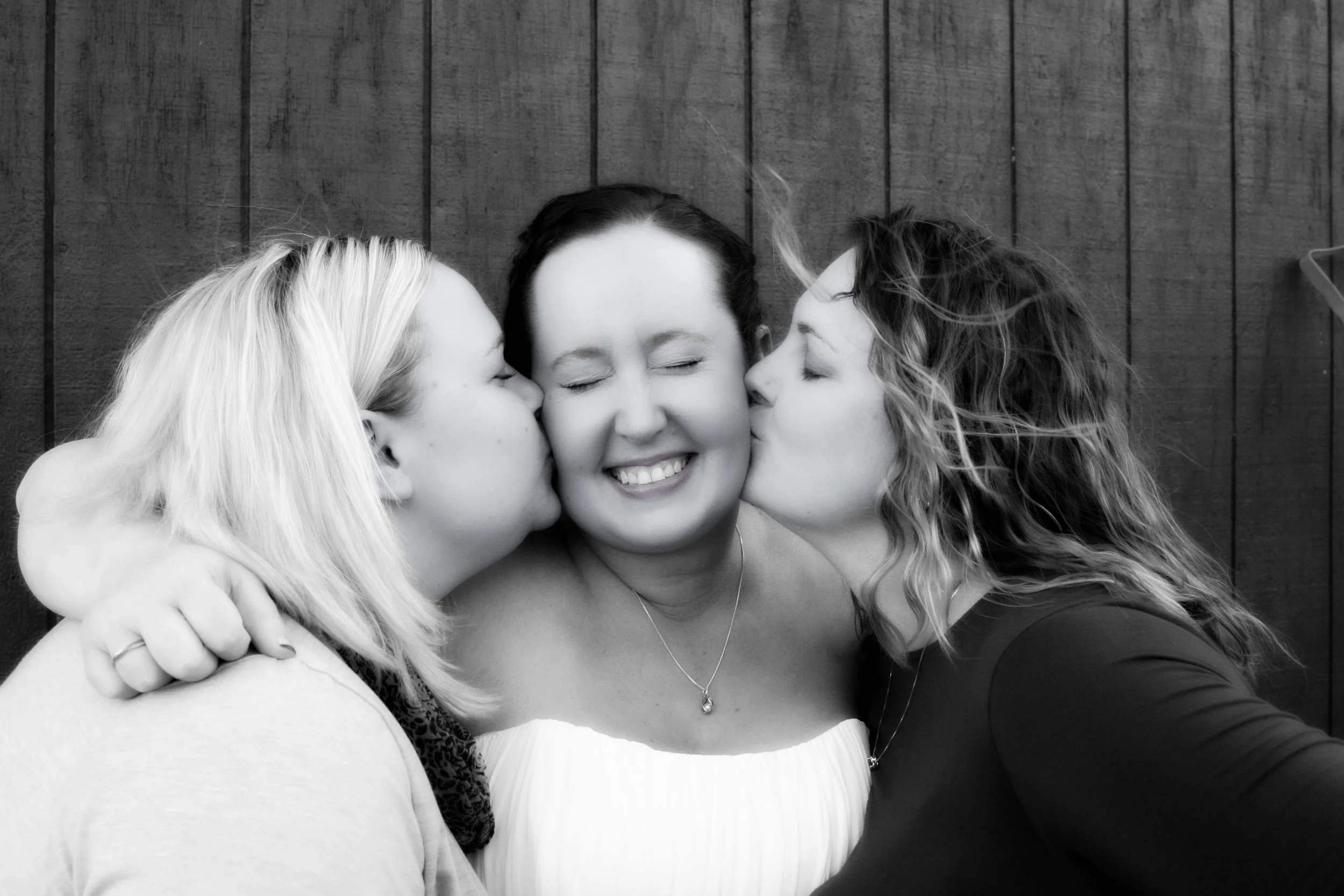 Three women, one in the middle with dark hair and two on the sides with light hair, sharing kisses on each cheek while smiling, against a wooden background.